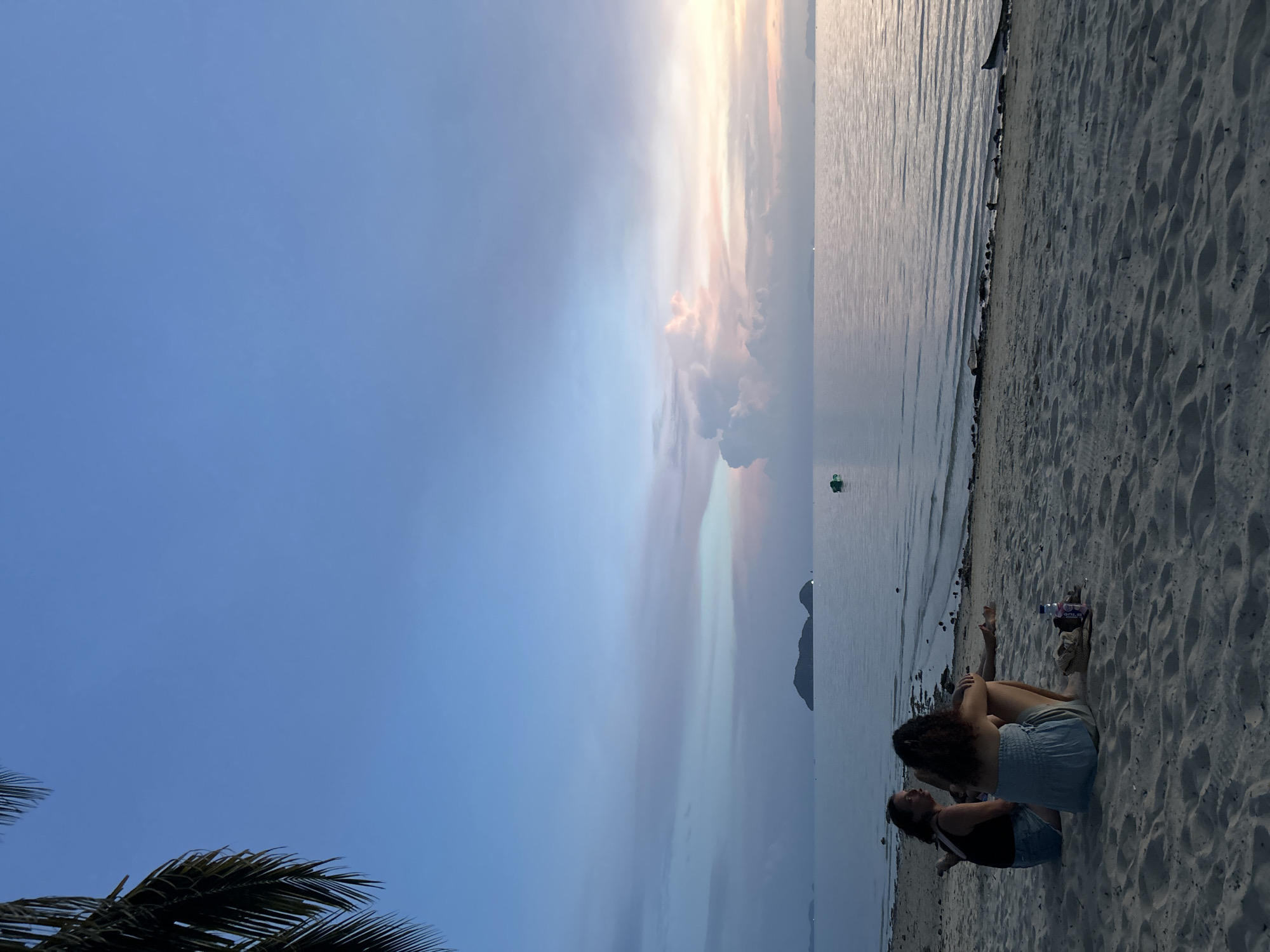 Two individuals are seated on a sandy beach, facing the calm ocean. The sky is a gradient of blues and soft pinks, indicating either dawn or dusk. In the distance, silhouetted islands break the horizon of the water, and a small green buoy floats near one of them. The scene suggests a peaceful moment of relaxation on a tropical coastline, possibly in Ko Pha-ngan District, Thailand, known for its beautiful beaches and islands.