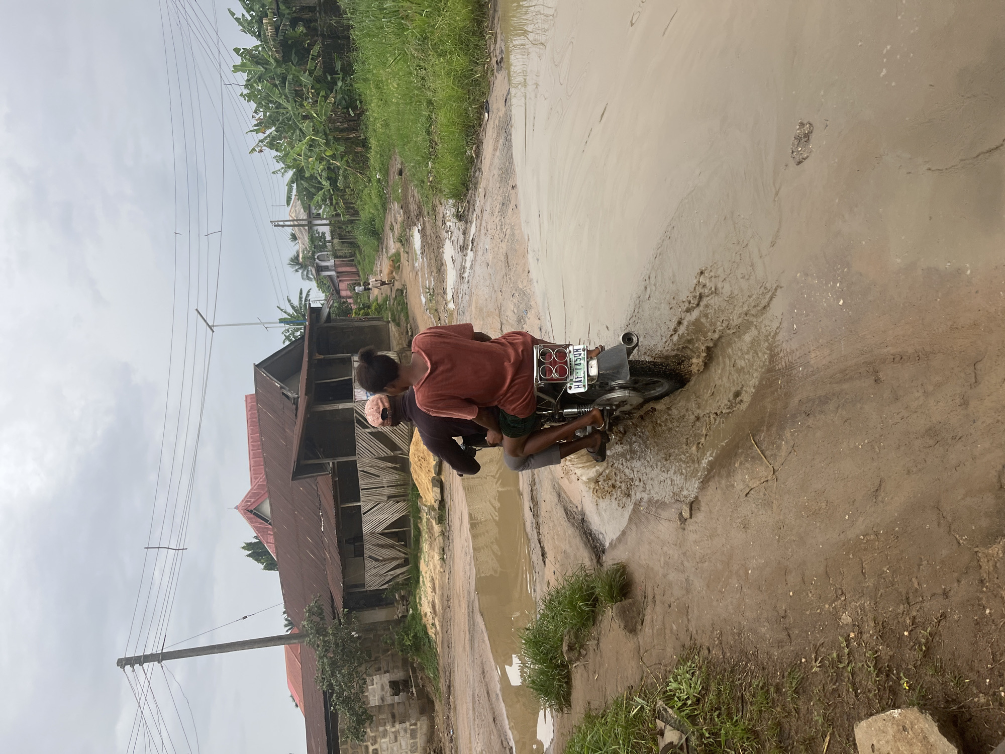 Two individuals on a motorcycle are traversing a waterlogged and muddy road in Ikot Ekpene, Nigeria. The road is heavily impacted by the weather, with large puddles covering the road. The buildings are under construction, with exposed building materials nearby. Overhead electrical lines are visible.
