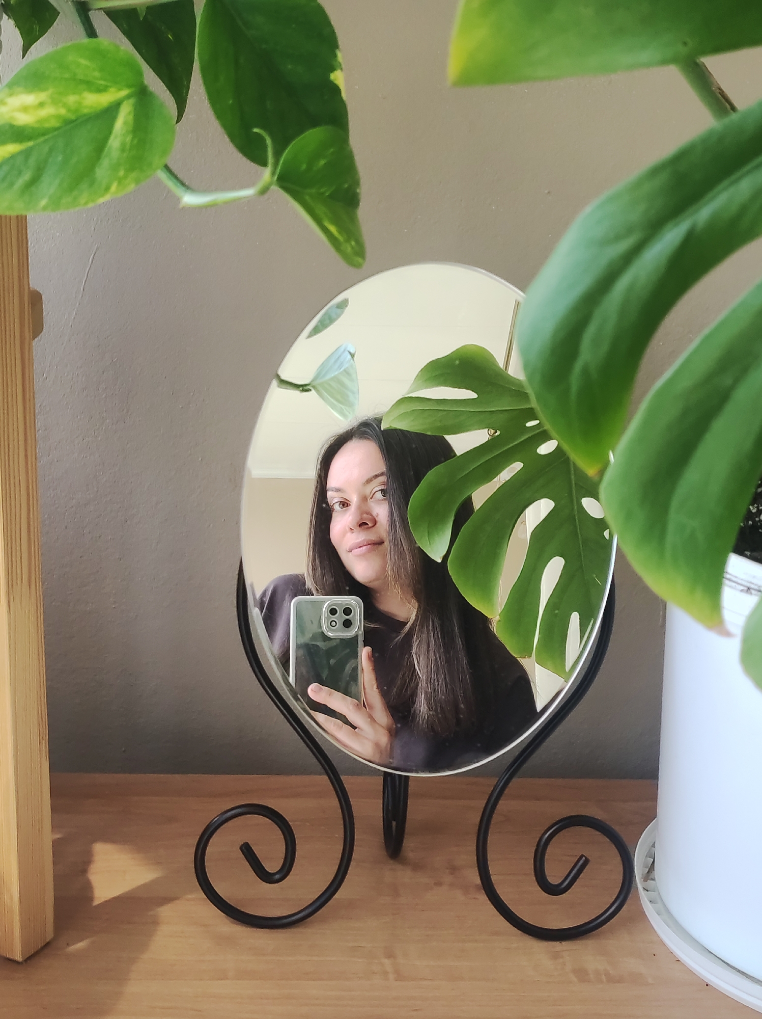 A medium close-up, indoor shot from Chaikovsky, Russia, captures a woman taking a mirror selfie. The woman, with long dark hair and a subtle, relaxed smile, is reflected in an oval-shaped mirror with a decorative black metal stand. She holds a smartphone, likely an iPhone given its distinct multi-lens camera array, to capture her reflection.
The mirror is positioned on a light-colored wooden surface, which could be a shelf or dresser. The scene is framed and accented by two distinct types of houseplants. In the upper left, variegated Pothos leaves with green and yellow patterns drape into the frame. On the right, a large, deeply lobed green leaf of a Monstera deliciosa plant partially obscures the mirror and a white plant pot, which also rests on the wooden surface. The background behind the mirror is a plain, light grey or beige wall. Natural light illuminates the setting, highlighting the plants and the woman's face, indicating it is daytime. No discernible text is visible in the image.