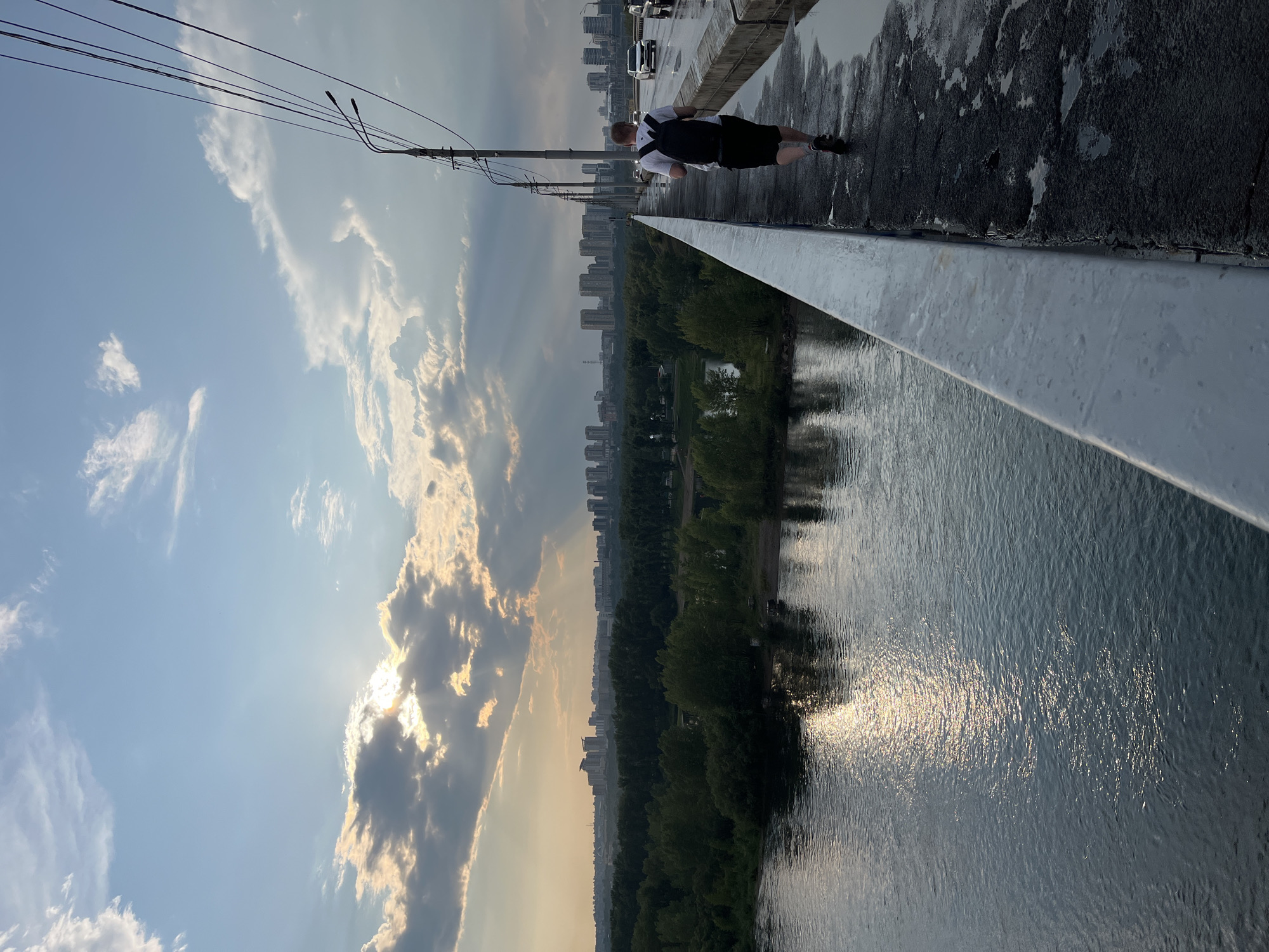 The image depicts a person walking on a bridge in Krasnoyarsk, Russia, with a panoramic view of the city and a river. The person, seen from behind, is wearing a white t-shirt and black shorts and carries a black backpack. They are walking on the right side of the bridge, on a wet asphalt path with puddles reflecting the sky. A white metal railing separates the path from the river. The river, which appears to be the Yenisei River, flows calmly below, with the sun's reflection creating a shimmering effect on its surface.
In the background, a dense line of green trees separates the river from the city skyline. The city features a cluster of modern high-rise buildings. The sky above is a mixture of blue and dramatic clouds, with rays of sunlight breaking through the clouds, suggesting either sunrise or sunset. The presence of power lines and streetlights on the bridge indicate it is a functional transportation route within the city. Several cars are visible on the bridge in the distance, heading in the same direction as the pedestrian. The overall atmosphere is one of a slightly overcast, yet visually striking, urban landscape.