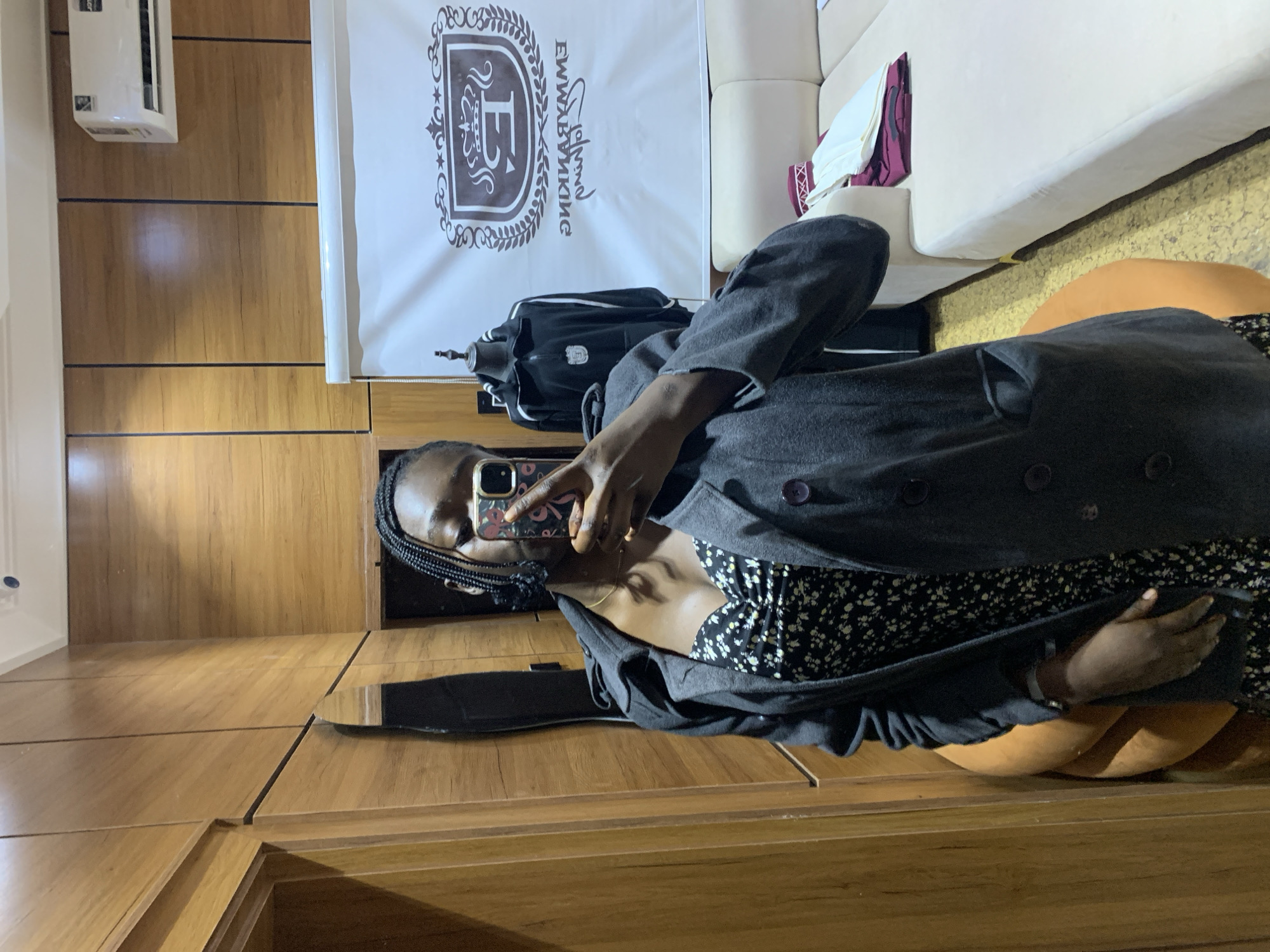 A young Black woman with braided hair takes a mirror selfie indoors. She is wearing a dark gray trench coat over a black and white floral print dress. Her right hand holds a phone with a floral case, and her left hand is visible near her hip. The background shows a room with wood-paneled walls, an air conditioning unit mounted high on the wall, and a white banner with a crest and text. A mannequin wearing a black athletic jacket stands behind her, and a cream-colored sofa is visible to the right. The location is identified as Gura Top, Nigeria.