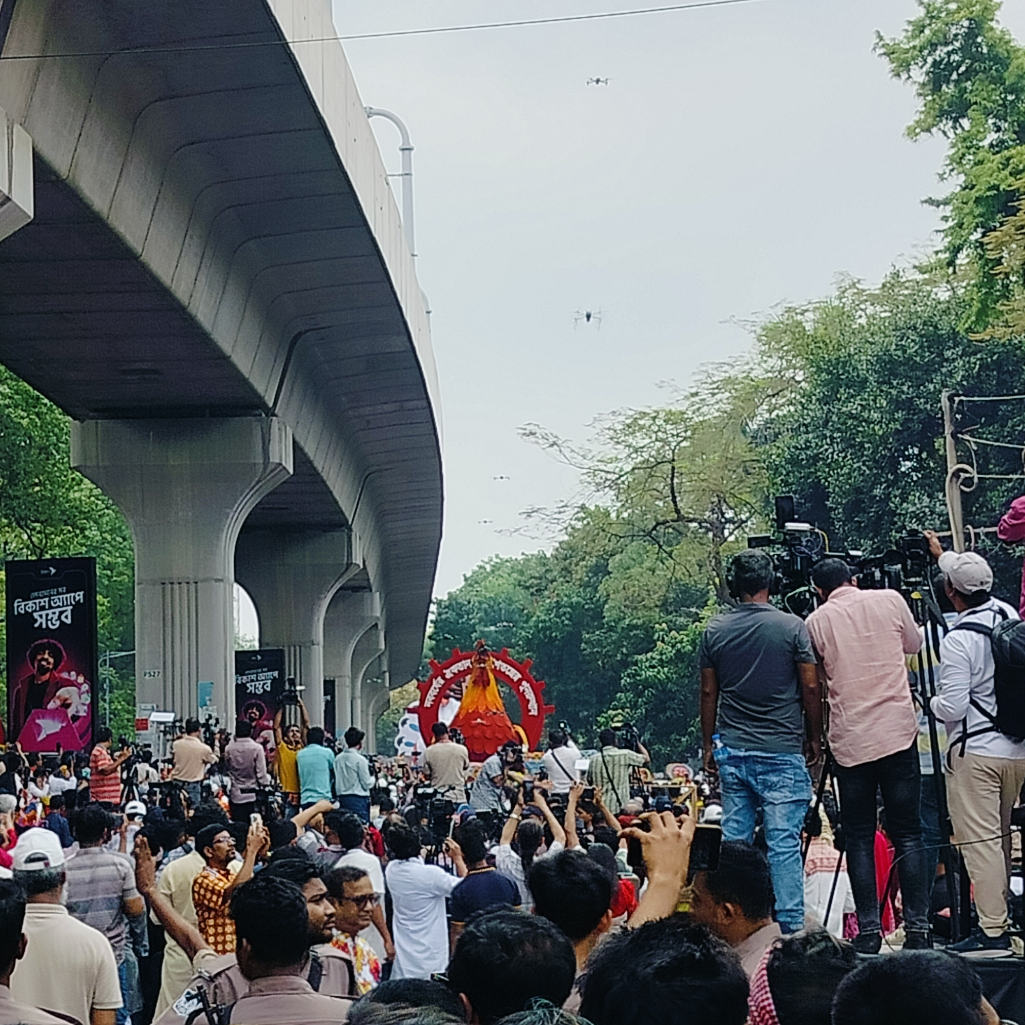 The image depicts a crowded street scene in Dhaka, Bangladesh, likely during a public event or gathering. A large group of people, primarily men, are visible, with many holding up phones to take pictures or videos. Several cameramen with professional video cameras are positioned on an elevated platform, recording the scene. In the center of the crowd, there is a large, ornate red and orange decorative display, possibly a symbol or centerpiece for the event. An elevated metro line structure looms overhead on the left side of the frame. Green trees line the right side of the frame. Several drones are visible in the sky above the crowd. Banners with Bengali script are attached to the metro line supports.
