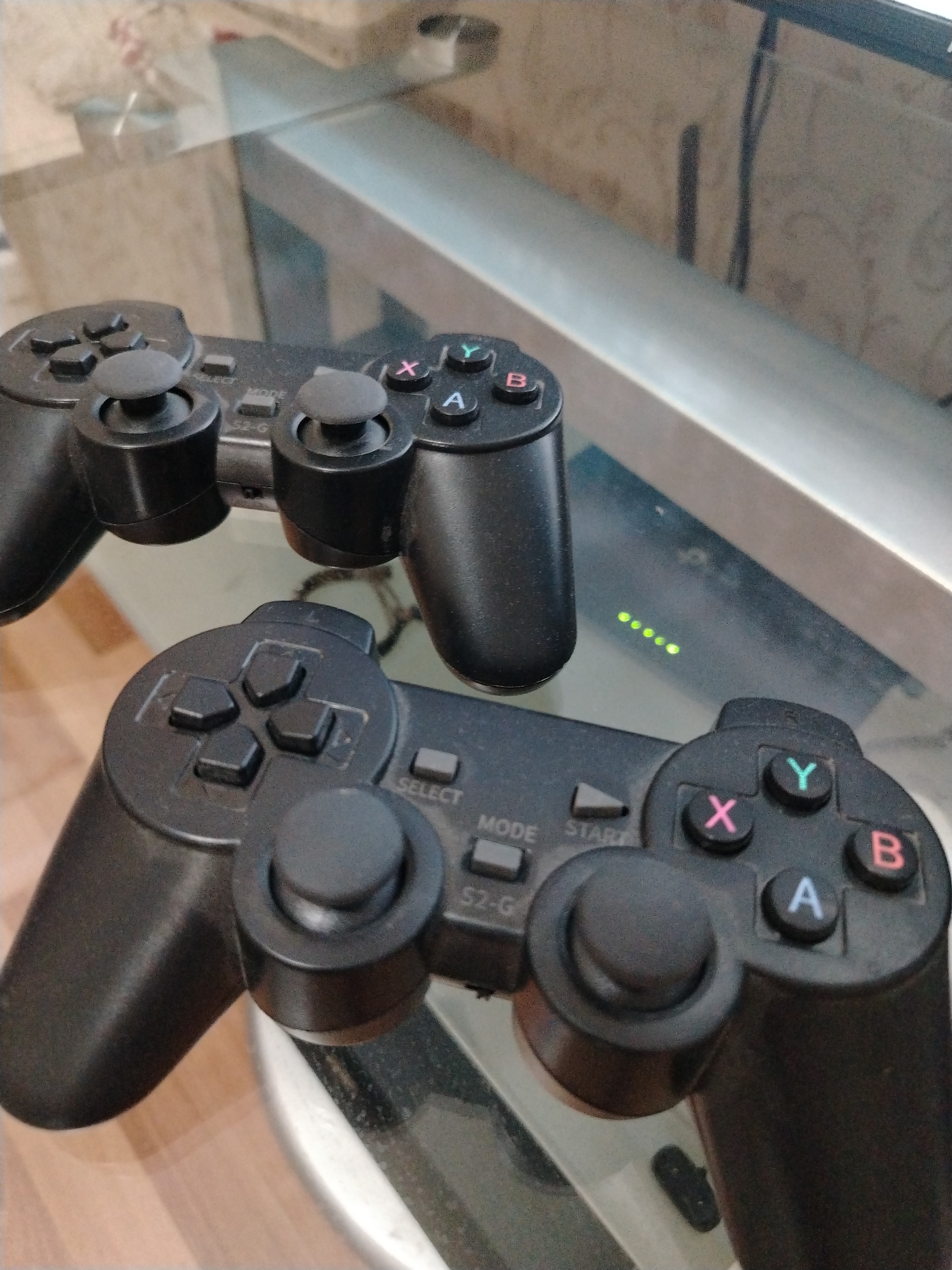 Two black video game controllers are placed on a glass table. The top controller is slightly angled and partially obscured by the bottom controller. The bottom controller is positioned horizontally and is more in focus. Both controllers have analog sticks, directional pads, and face buttons labeled with letters (X, Y, A, B) and symbols. Some buttons on the bottom controller are visible, including "SELECT," "MODE," and "START." To the right of the bottom controller, on the surface beneath the glass, are several green LED lights arranged in a horizontal line. The background of the image is blurred and appears to be an indoor setting with some reflective surfaces and a wooden floor. The image does not contain any identifiable landmarks or elements that would suggest a specific location within Krasnodar, Russia.