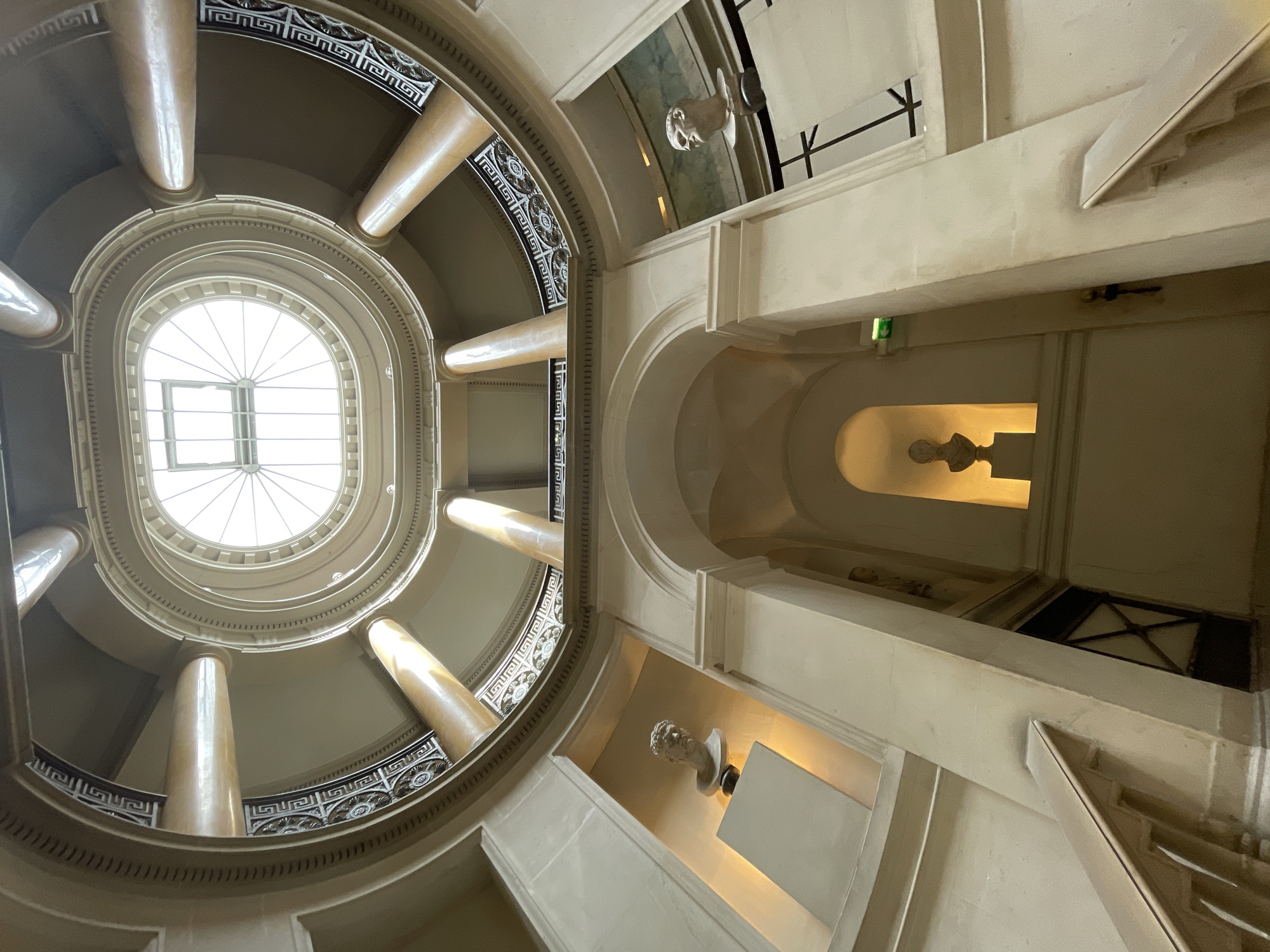 The image shows the interior of a rotunda in Paris, France. The rotunda has multiple levels, with columns and balconies visible. A skylight at the top illuminates the space. Two busts of historical figures are displayed on pedestals on different levels. One bust is on the left side, and the other is on the right side. There are staircases leading up through the rotunda. The architecture is classical, with ornate detailing on the railings and circular elements. The lighting is warm and diffused, suggesting an interior setting.