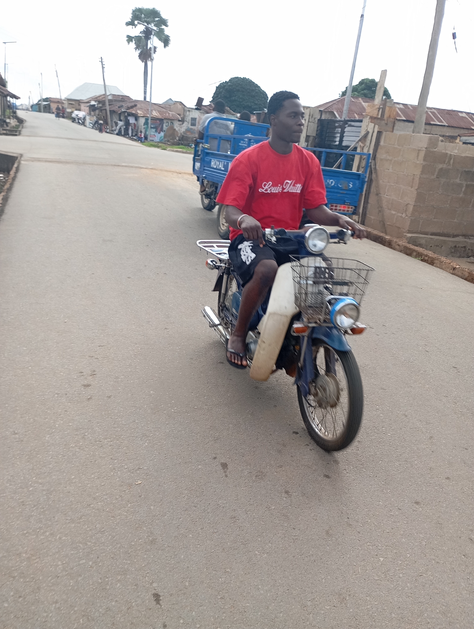 A man wearing a red t-shirt with "Louis Vuitton" printed in white text and black shorts is riding a blue motorcycle with a white front fender and a basket on the handlebars. The motorcycle has a license plate that reads "ROYAL". The man is wearing black flip-flops. The scene takes place on a paved road in Shendam, Nigeria. In the background, there are buildings with corrugated metal roofs, a blue truck with a similar "ROYAL" logo, and a tall palm tree. The sky is overcast. The man is looking to his left, and his hands are on the handlebars. The motorcycle's front wheel is in motion.