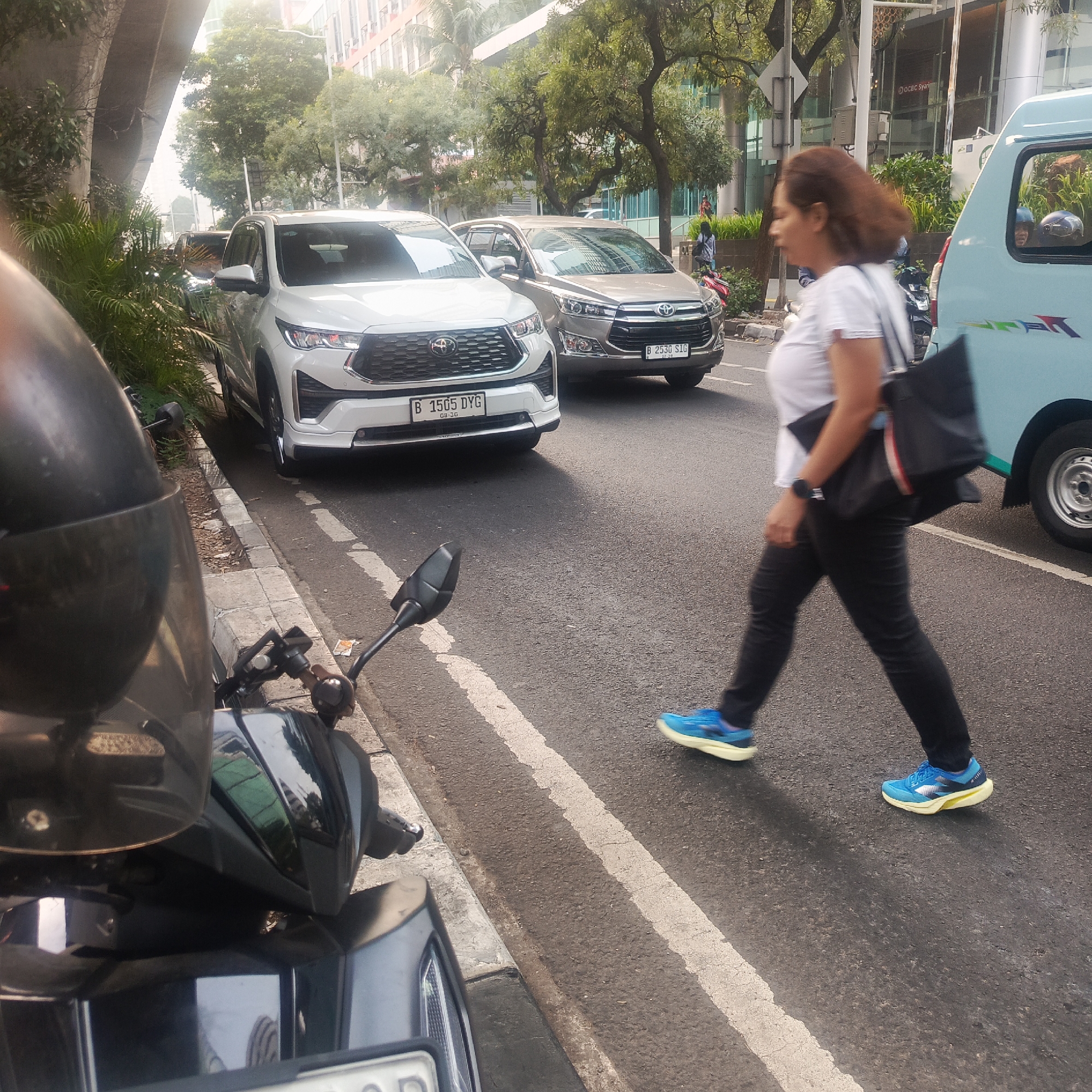 In South Jakarta, Indonesia, on a paved road with a white line marking a pedestrian crossing, a woman wearing a white shirt, black pants, and blue athletic shoes is walking from left to right across the street. She is carrying a black tote bag over her right shoulder. In the background, a white SUV, identified by its license plate "B 1505 DYG," and a silver SUV with license plate "B 2530 SIG" are stopped on the road. To the right of the SUVs, a light blue van is also present. A motorcycle is partially visible in the foreground on the left side of the image. The environment includes trees and buildings, suggesting an urban setting.