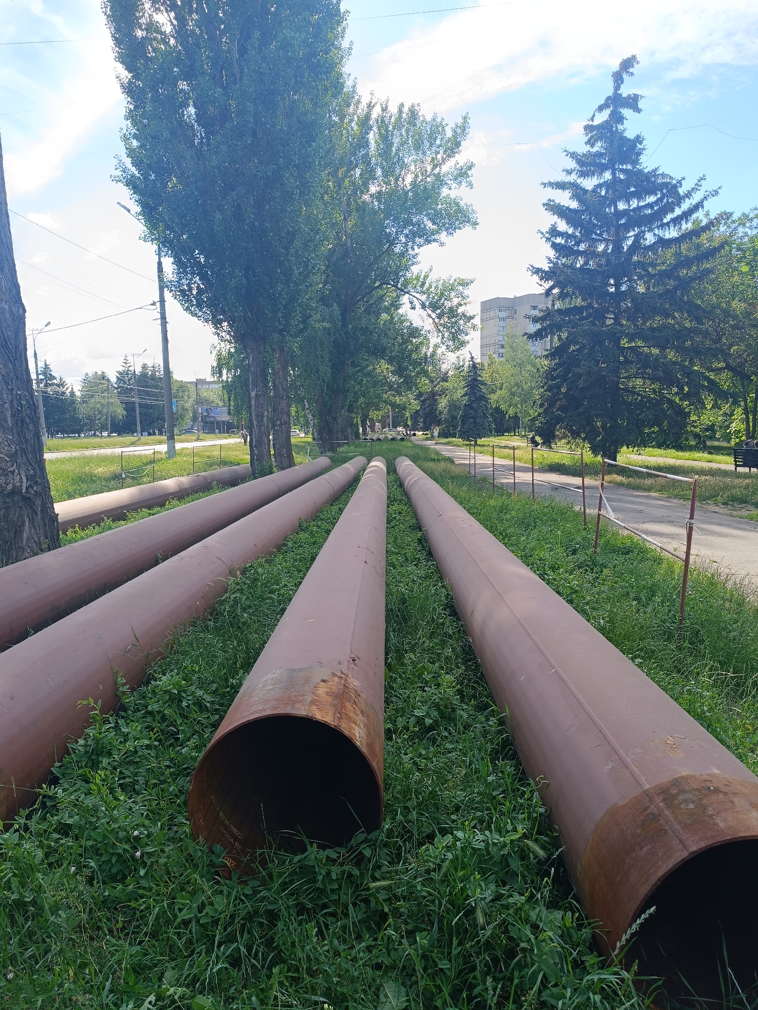 The image depicts a sunny day scene along a residential street in Kryvyi Rih, Ukraine. In the foreground, several large, rusty, brown pipes lie parallel to each other on a grassy area next to a paved walkway. The pipes are spaced apart and appear to be laid out for construction or maintenance purposes.
Beyond the pipes, a walkway extends into the distance, bordered by a low fence with white and red posts. Trees line the walkway on both sides, with tall, slender trees with green foliage on the left and a dense, dark green evergreen tree on the right. Further in the background, a multi-story apartment building is visible, interspersed with more trees. The sky is blue with scattered white clouds. Two people are visible sitting on benches on the walkway in the middle ground. The environment appears to be a well-maintained urban park or residential area.