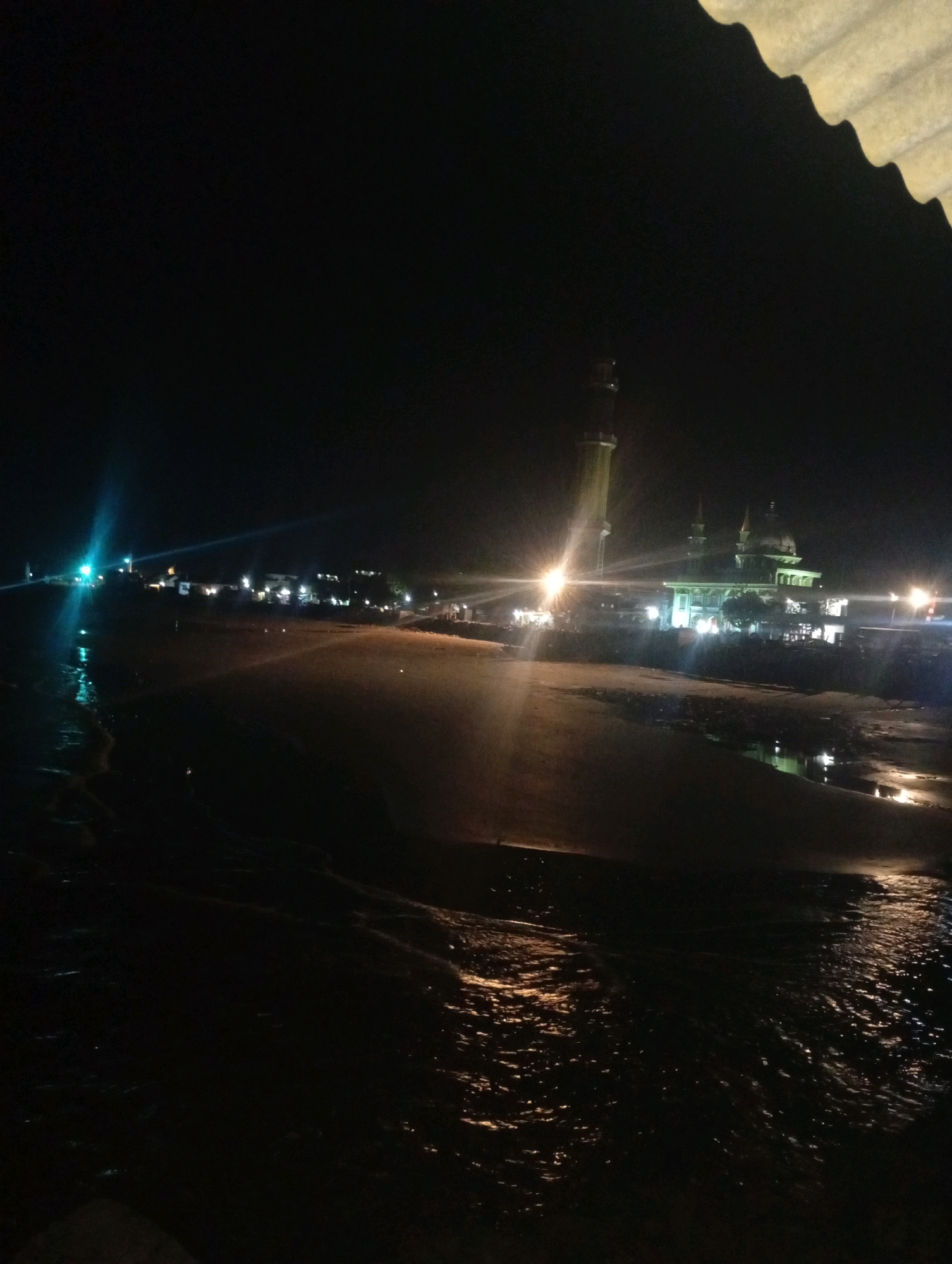 The image depicts a night scene on a beach near a mosque in Banowan, Indonesia. The foreground shows dark, wet sand and rippling water reflecting artificial lights from the shore. In the midground, a sandy beach stretches towards a row of buildings illuminated by lights. Prominently featured is a mosque with a tall minaret and domed structures, brightly lit. Several other buildings, likely shops or residences, are visible along the coast, their windows and lights creating a scattered glow against the dark background. The overall atmosphere is tranquil, with the sounds of gentle waves implied by the water's movement. No people are clearly visible in the frame. The sky is completely dark, suggesting it is late night or early morning.