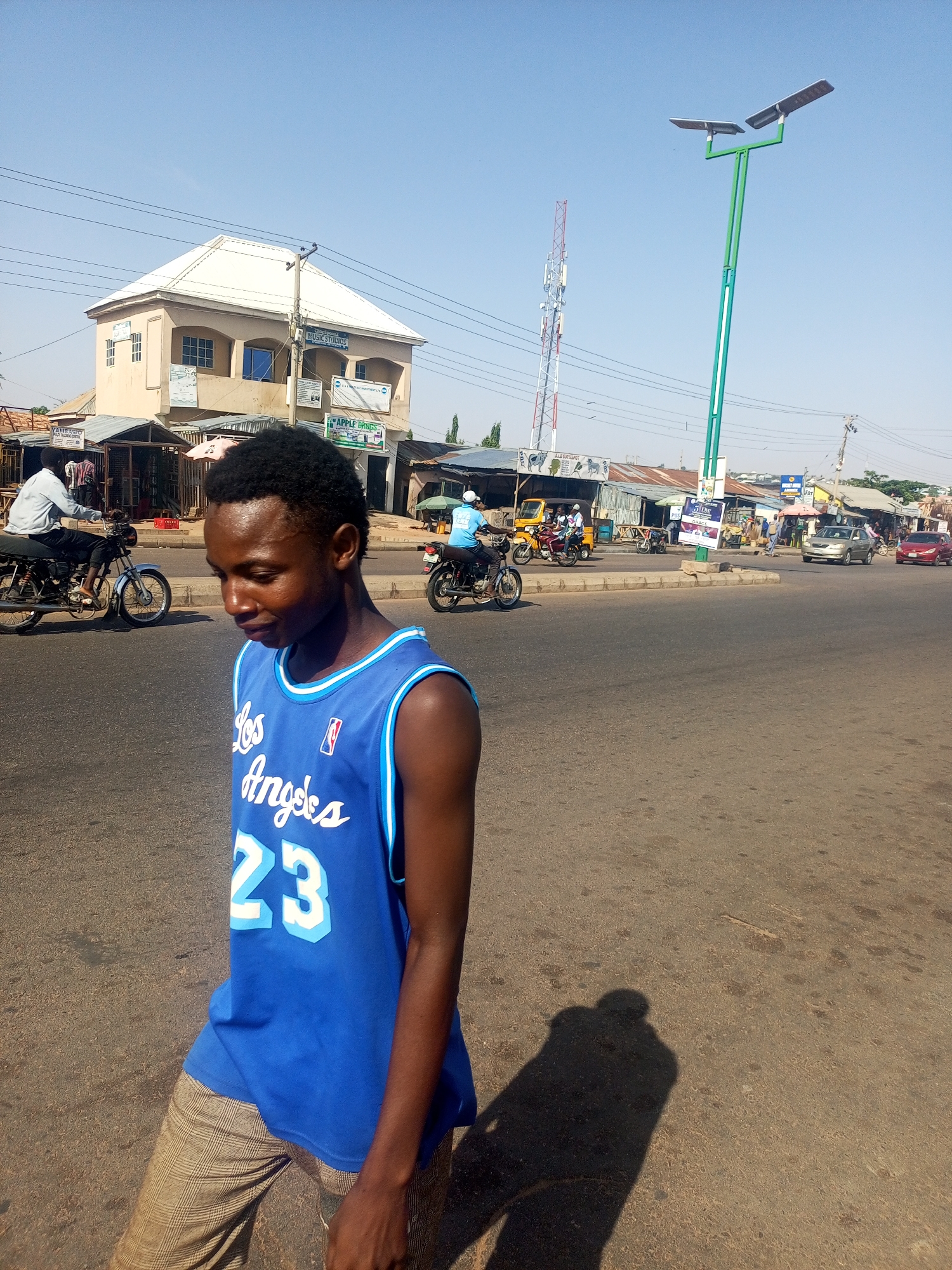 This daylight photograph captures a vibrant street scene in Tempure, Nigeria, under a clear blue sky. The main subject is a young Black man in the foreground, walking towards the left with his head tilted down. He is wearing a blue Los Angeles Lakers basketball jersey with "Los Angeles" and "23" printed on the front in white, and light brown plaid pants. His long shadow is cast on the asphalt road to his right, indicating bright sunlight, likely mid-morning or late afternoon.
In the mid-ground, a two-story building with a light-colored roof houses multiple businesses, including "MUSIC STUDIOS," "P.K.L.D INVESTMENT LTD," and "APPLE DRUGS." Below it are several market stalls with corrugated iron roofs, one identified as "YAME AUTO" and another as "ALL RITA SHOP." The wide, paved street is busy with various forms of transportation, including several motorcycles (some with riders), a gold sedan, a red car, and a three-wheeled Keke Napep.
Prominently featured on the right side of the street is a tall, modern green streetlight equipped with two solar panels. In the background, a red and white telecommunications tower stands tall against the horizon. Overhead power lines crisscross the sky. The overall scene depicts a lively urban or semi-urban area characterized by local businesses, active road traffic, and typical infrastructure.