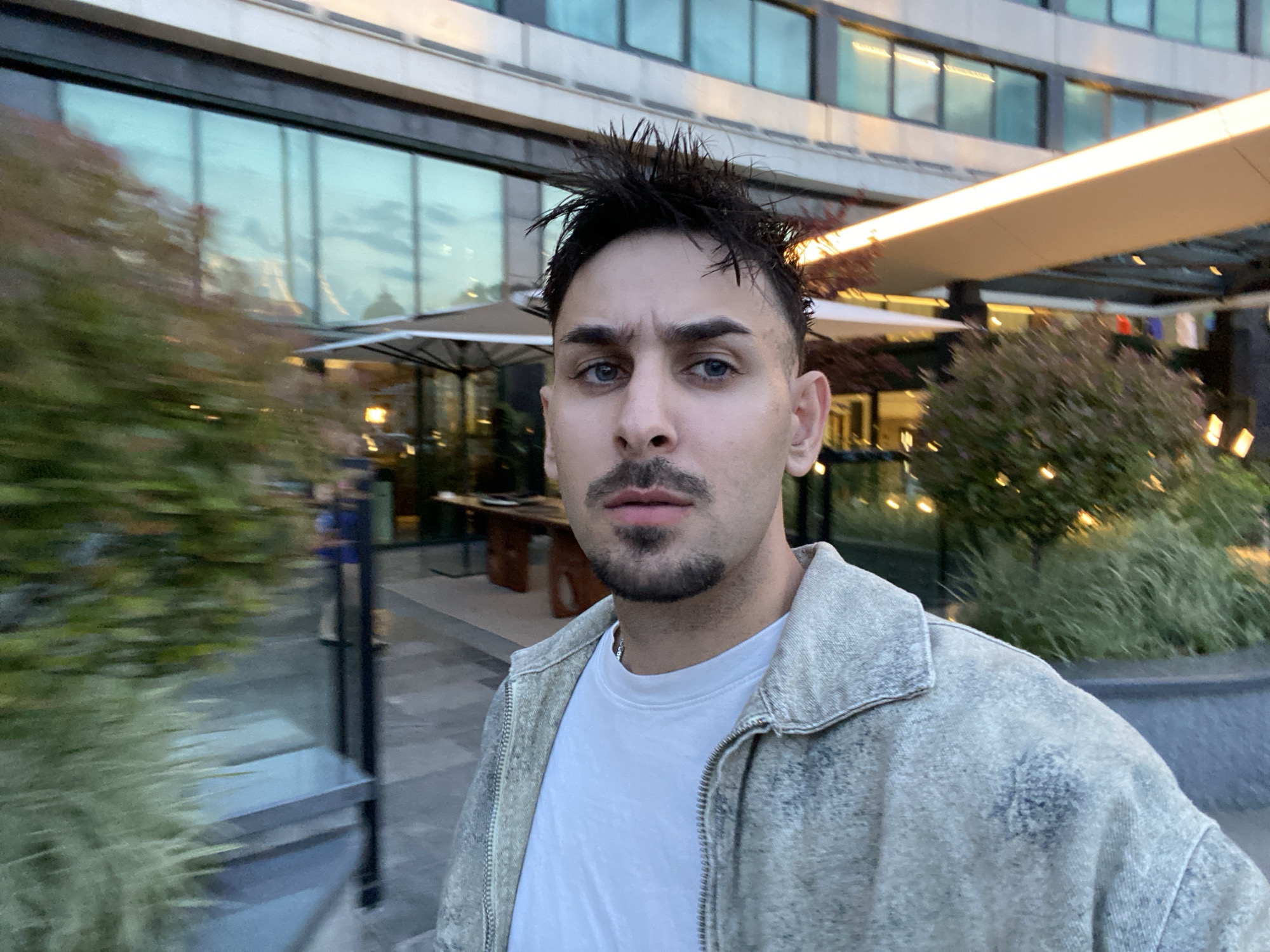 A young man, facing the camera in what appears to be a selfie, is positioned outdoors in Sofia, Bulgaria. The location is identifiable as the area surrounding the Millennium Center. He has styled dark hair, prominent eyebrows, blue eyes, a mustache, and a goatee beard, and maintains a neutral expression. He is dressed in a white t-shirt layered under a light-colored, possibly acid-wash or distressed, zipped jacket.
Behind him, the modern facade of the Millennium Center building rises, characterized by multiple floors of large, reflective glass windows that display varying hues of blue, green, and reflections of the sky and adjacent structures. An illuminated horizontal canopy extends from the building over an outdoor area, where white umbrellas and tables suggest a cafe or restaurant. Lush green bushes and foliage are visible on both sides of the frame, with some on the left exhibiting motion blur, hinting at either camera movement or a shallow depth of field. The scene is bathed in the soft light of what appears to be late afternoon or early evening, complemented by subtle artificial lighting from the building and outdoor amenities. The ground is a paved urban surface.