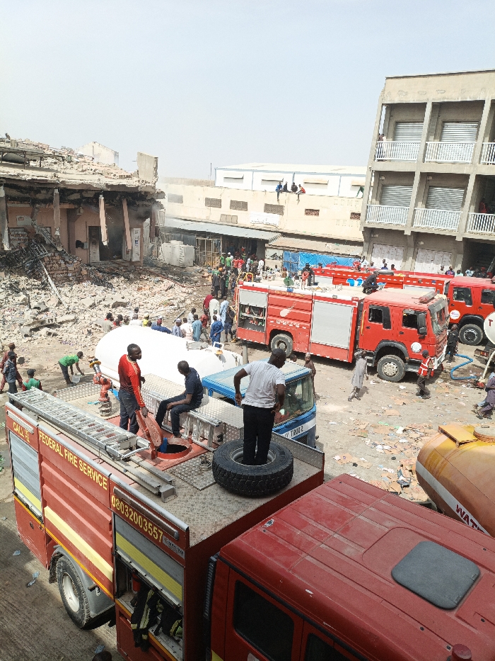 The image displays an emergency response scene in Fagge C, Nigeria. A red Federal Fire Service truck, labeled with "CALL 444" and "08032003557," occupies the foreground, its roof serving as an observation point for three individuals: one man in a white t-shirt standing on a tire, another in a black uniform seated near an open circular hatch, and a third in a red and black shirt standing beside a different access panel.
Immediately behind this truck, a blue and white tanker truck with a large white tank is positioned, surrounded by several individuals, one in a green shirt appearing to interact with equipment. The ground in the mid-ground is dusty and strewn with debris, paper, and scattered refuse.
To the left, a multi-story building exhibits extensive damage, with its upper floors collapsed into a large pile of rubble, exposing rebar and structural components. Charring is visible within the remnants, suggesting fire damage. To the right, a multi-story building with multiple balconies and a uniform facade appears structurally intact.
In the mid-ground, at least two additional red fire trucks are parked. Numerous individuals, appearing to be a mix of emergency personnel and civilians, are dispersed throughout the scene, observing and engaging in various activities near the vehicles and the damaged structure. Several people are visible seated on the roof or ledge of a lower building in the background, observing the incident. The scene is illuminated by clear daylight, indicating an outdoor, daytime event.