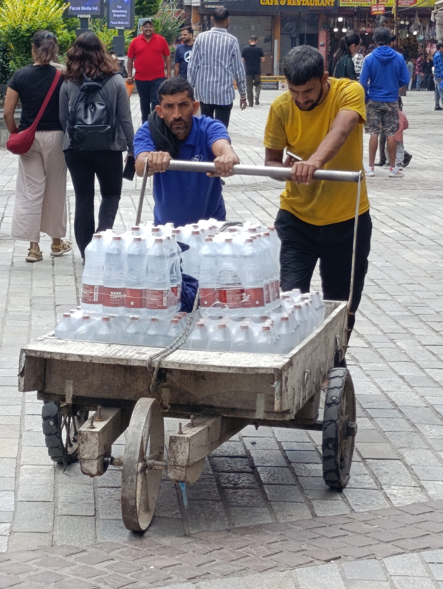 Two men are pushing a cart loaded with bottles of water down a cobbled street in Manali, India. The man on the left is wearing a blue shirt and the man on the right is wearing a yellow shirt. Both men are pushing the cart from behind. The cart is old and made of wood and metal. It has two large wheels at the back and two smaller wheels at the front. The bottles of water are stacked high on the cart, covered in plastic wrap. In the background, other people are walking and shops are visible. The street is wet, suggesting it may have recently rained. The overall scene depicts a common street vendor scene in a busy town.