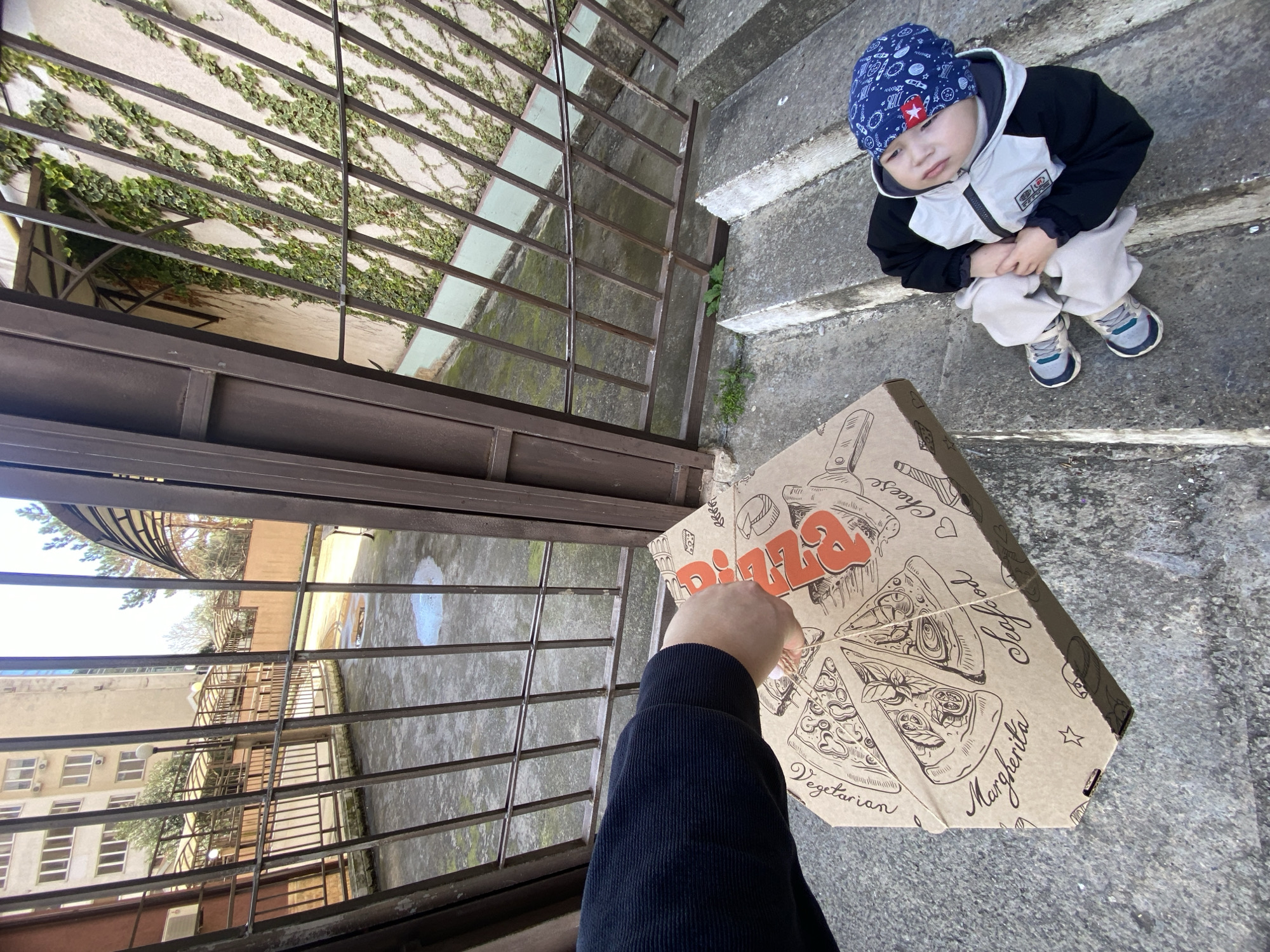 A young child, wearing a blue hat with a star, a black and white jacket, and light-colored pants, sits on concrete steps. A pizza box is being handed to the child through a metal gate. The building behind the gate appears to be a multi-story structure. The location is likely Sochi, Russia.