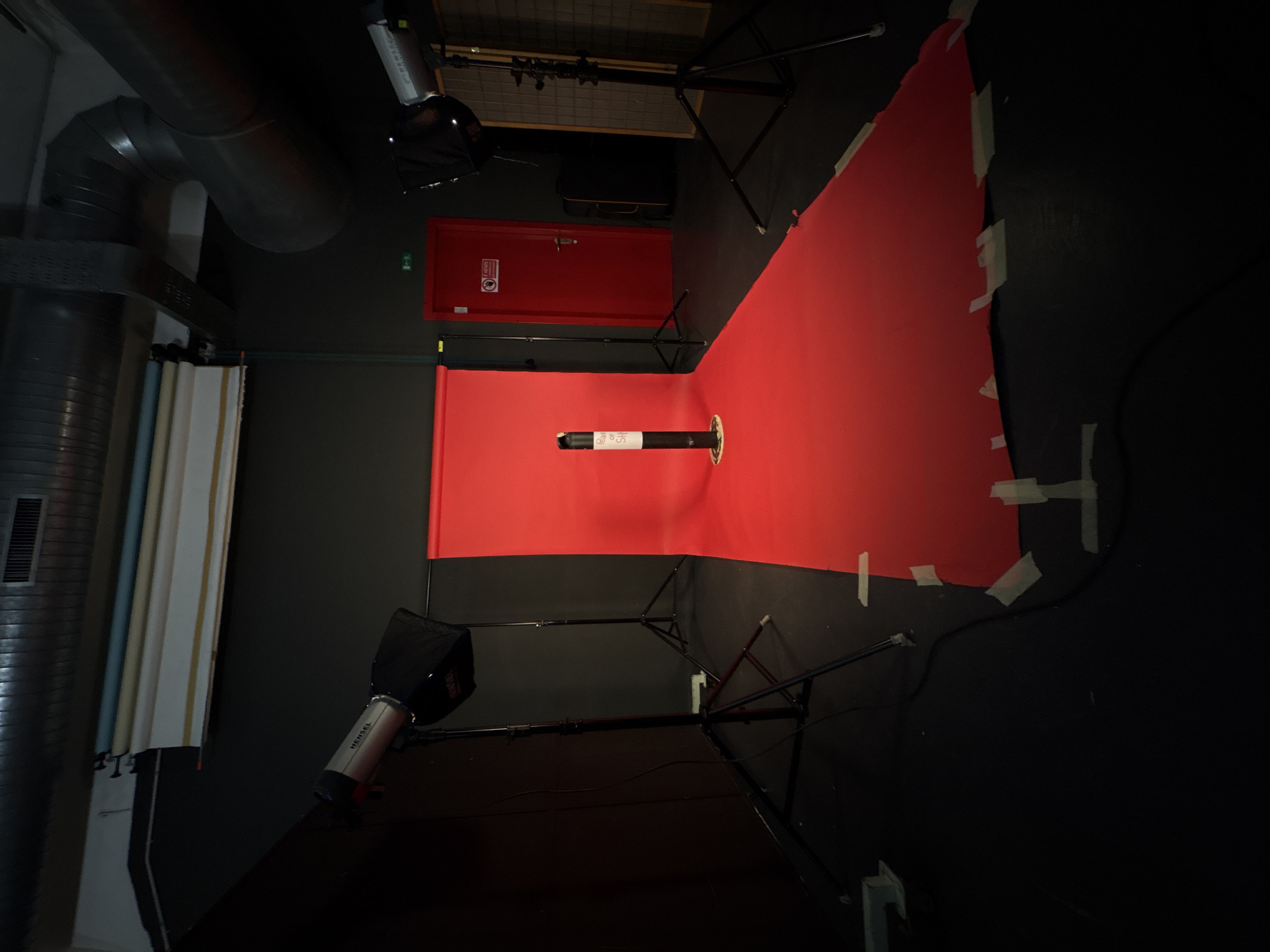 The image captures an indoor studio environment, likely set up for photography or videography. The scene is dominated by a vibrant red seamless backdrop, meticulously taped down to a dark floor and extending up a dark wall, creating a continuous background.
**Objects and Equipment:** Two studio lights, one clearly branded "HENSEL," are positioned on stands with modifiers (possibly softboxes), indicating professional lighting equipment. One is in the foreground bottom left, and another is in the background top right. Against the left wall, several rolls of various colored backdrops are stored vertically. In the center of the red backdrop stands a black cylindrical object with a white label that reads "PRO OF SH." Large, grey industrial-style air ducts are visible in the upper left, running along the ceiling. A red door with a white-on-red fire safety sign (partially legible, including "NOTFALL" and other text) is in the background. A black equipment case is visible on the floor near the back right.
**Scene and Setting:** The walls and floor are dark, suggesting a controlled environment designed to minimize reflections and outside light. The overall atmosphere is functional and prepared.
**Activity:** The setup suggests that a photoshoot or video recording is either about to take place or has recently concluded, utilizing the red backdrop and studio lighting.
**Notable Details:** The clean, seamless appearance of the red backdrop and the careful placement of equipment highlight the intentional nature of the space. The industrial elements contrast with the controlled studio setup.