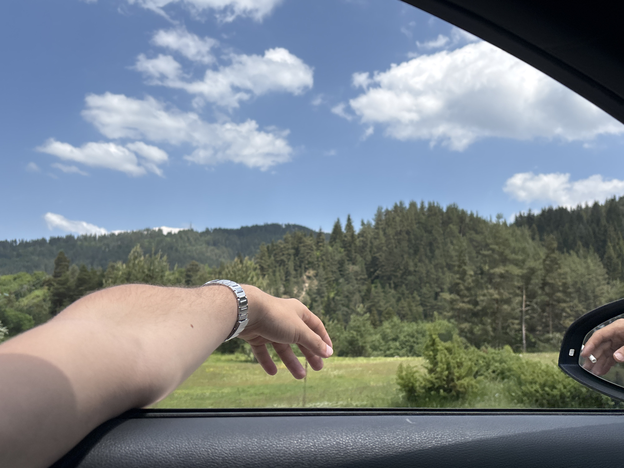 The image depicts a rural mountainous landscape viewed from the open passenger-side window of a vehicle, in the vicinity of Beli Iskar, Bulgaria. A human right forearm and hand, fair-skinned and lightly haired, extends outward from the window. The elbow rests on the textured black interior trim of the car's door panel. A silver-colored metal link bracelet or watch is present on the wrist, and a small, dark mole is visible on the upper forearm near the elbow. The hand is open, with fingers slightly curled downwards. A partial reflection of the hand and bracelet is visible in the car's right-side mirror.

Outside the vehicle, a vibrant green field or meadow occupies the midground. Beyond the meadow, a dense forest of coniferous trees covers rolling hills and mountains, forming the background. The trees are uniformly green and appear robust. The sky above is clear blue with scattered, fluffy white cumulus clouds, indicating bright daylight conditions.