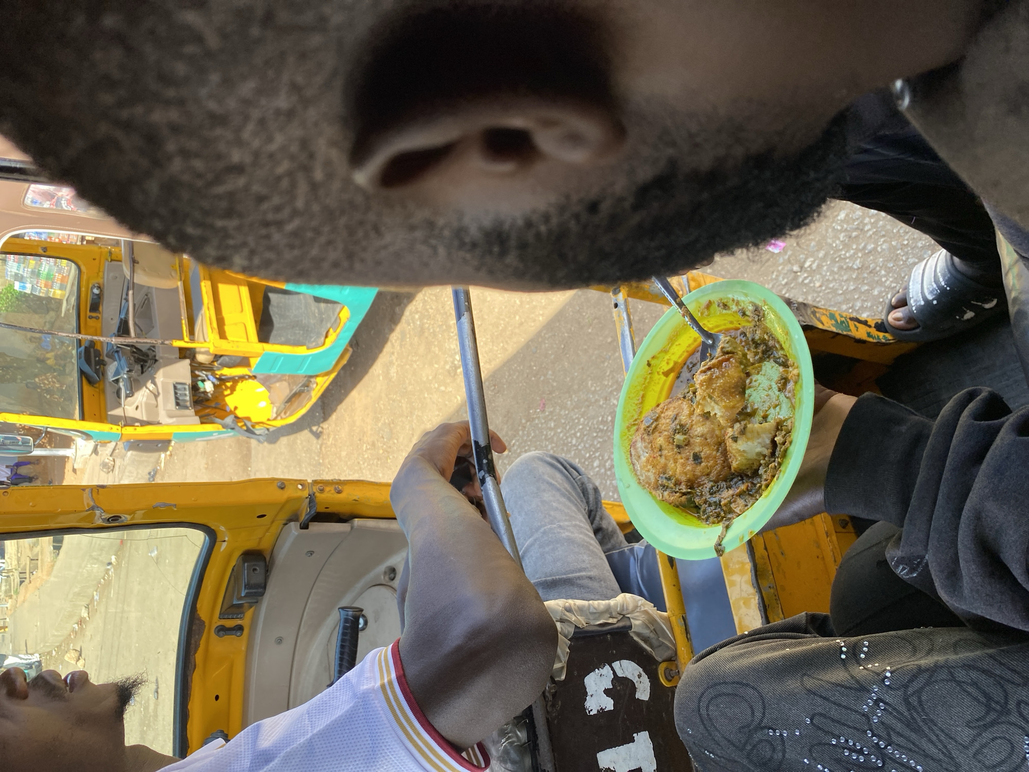 The image provides a first-person perspective from inside a yellow auto-rickshaw, likely a Keke NAPEP, in Bukuru, Nigeria, under bright daylight conditions. The frame is dominated by the chin, lower lip, and beard of an individual in the immediate foreground. This person's right hand holds up a bright green plastic plate containing a meal, possibly Akara (bean cakes) or fried plantains with a stew/sauce, with a spoon inserted into the food. They are wearing a dark long-sleeved top, dark pants with sparkling embellishments, and a black slide sandal on their visible foot.
To the lower left, another individual is partially visible, looking upwards. This person wears a white shirt with red and gold stripes and has a short beard. Their right arm rests on a silver bar, part of the vehicle's frame. The yellow interior and exterior of the auto-rickshaw are clearly visible, including seats and a window reflecting the sunny surroundings. Shadows cast by the vehicle and individuals indicate a bright, clear day. On a brown panel below the left person's arm, the white text "C1" is visible. The scene captures a casual moment of eating or preparing to eat while traveling or stationary in the local transport.