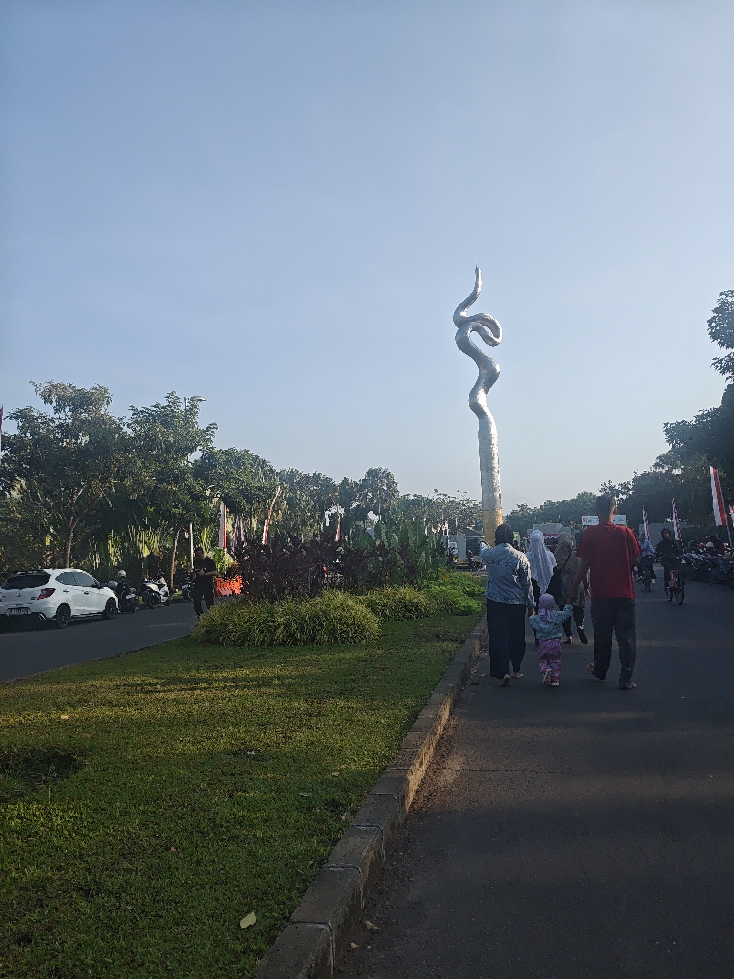 The image shows a public area with a large, abstract sculpture resembling a coiled snake reaching towards the sky. Several people are walking on a paved path adjacent to a grassy median strip. On the other side of the median, there is a road with a white car and several motorcycles parked. Tall trees and foliage line the background, with some flags visible among them. The sky is clear and blue, suggesting it is daytime. The scene appears to be in Bacin, Indonesia, a location known for its public art and green spaces.