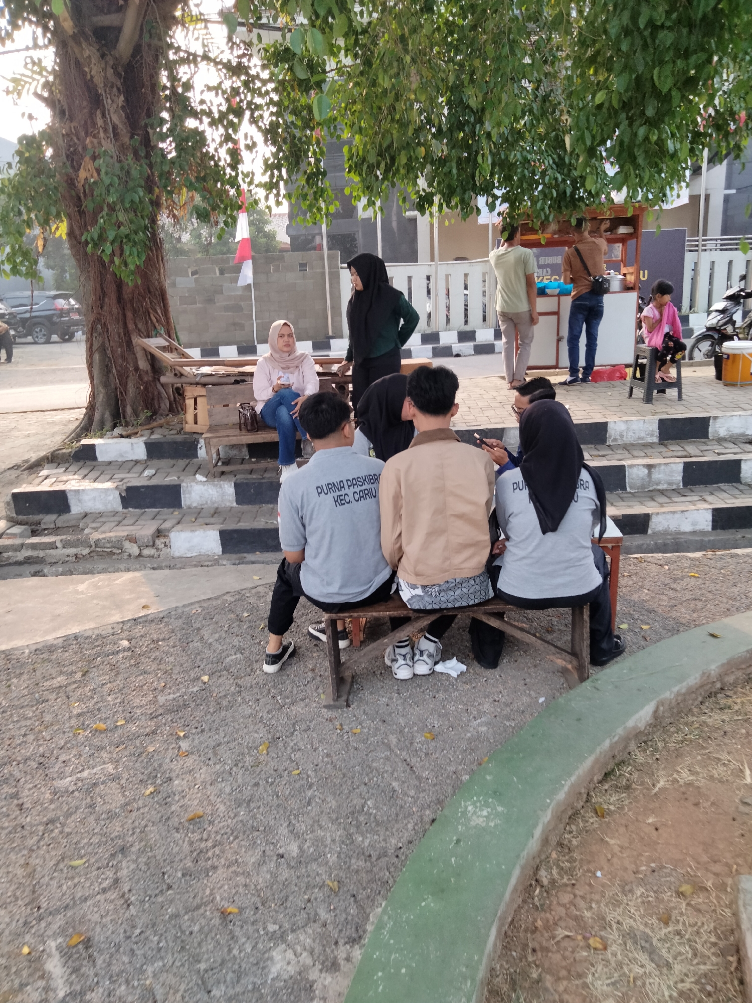 The image depicts an outdoor scene in Cariu, Indonesia, with a group of people gathered near a large tree. In the foreground, four individuals are seated on a wooden bench, facing away from the camera. The person on the far left wears a grey t-shirt with text that appears to read "PURA PASIKIBRIA KEC. CARIU". Next to them is another person in a tan jacket. To the right, two individuals are seated, both wearing headscarves. One is in a grey t-shirt and black pants, while the other is in a black headscarf and grey t-shirt.
In the background, a food cart is visible, with two men standing near it. One is wearing a light green t-shirt and tan pants, and the other is in a brown shirt and jeans, with a black bag slung over his shoulder. A young girl in a pink outfit is seated on a black stool to the right of the food cart. There are several motorcycles parked behind the food cart and a building with white railings in the distance.
The environment features a large tree with extensive roots and leafy branches. Steps with black and white markings are visible to the left of the bench where the first group is seated. The ground is covered in gravel and dry leaves. A curved green planter is in the lower right corner of the frame. The lighting suggests it is late afternoon, with sunlight filtering through the tree leaves.