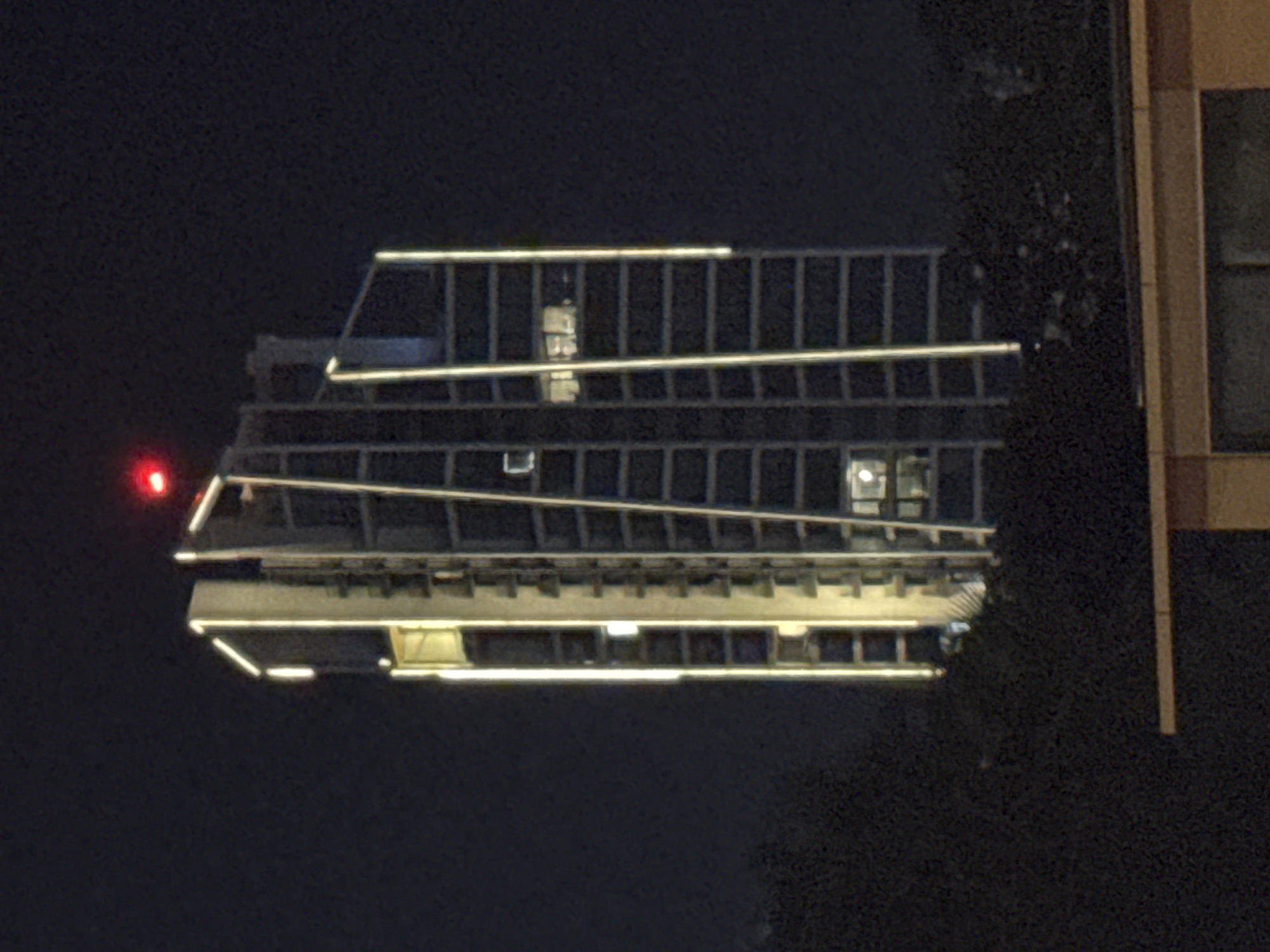 The image shows a tall, modern building at night in the Mouttagiaka Community, Cyprus. The building is illuminated by vertical lines of light along its facade and by lights within its numerous windows. At the very top of the building, a small red light is visible, likely a beacon for aviation. The building is surrounded by dark foliage, suggesting it is situated in an area with trees or a park. In the foreground, the top of another, lower structure is visible, with a light-colored band across its roof. The overall impression is of a contemporary urban or semi-urban environment at night.