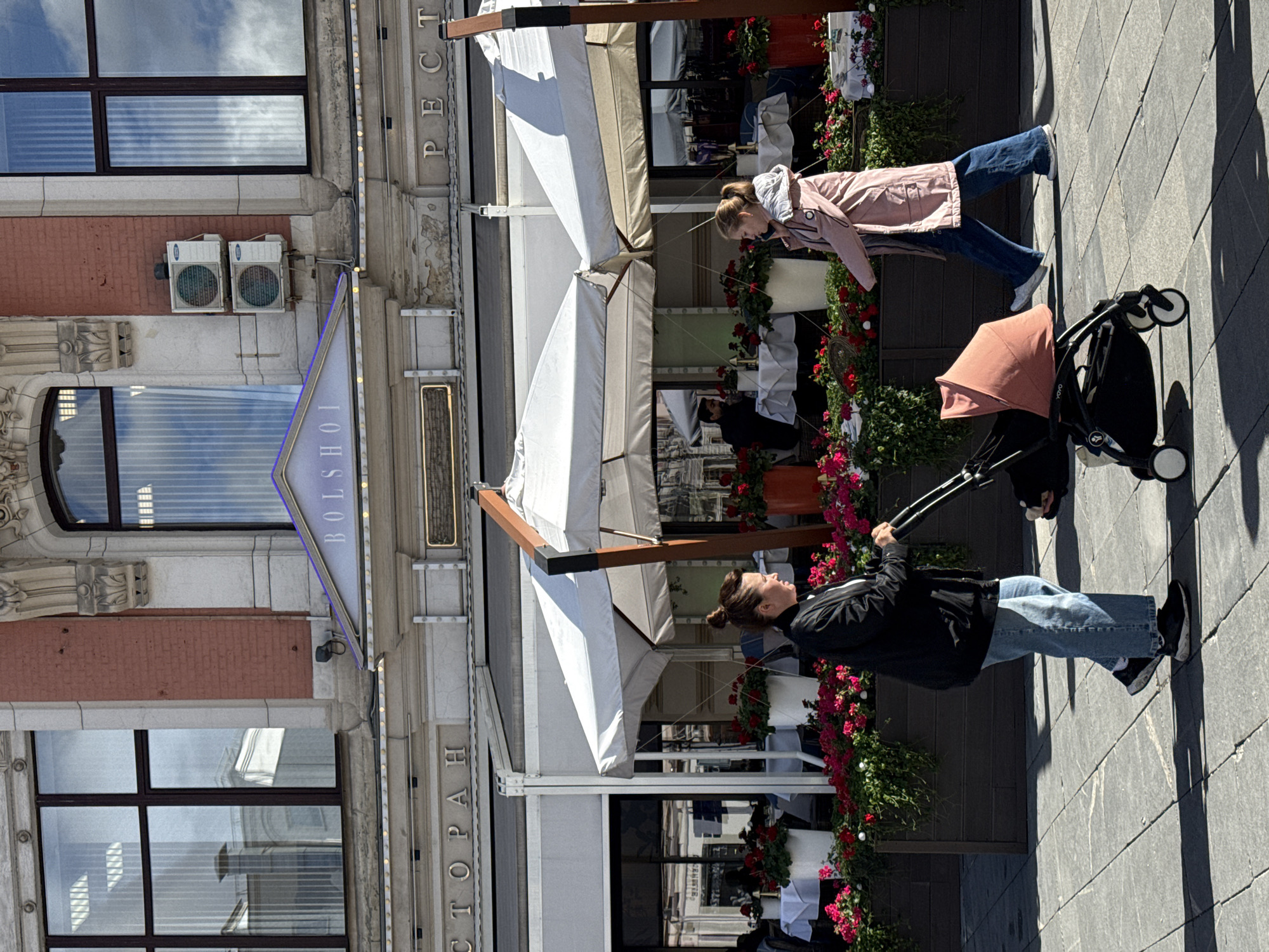 The image shows two women walking past a restaurant in Moscow, Russia. One woman is wearing a black jacket and jeans, pushing a stroller. The other woman is wearing a pink jacket and jeans, bending down to look at something. The restaurant has white awnings, red flowers in pots, and the sign "BOLSHOI." The building is brick and stone with large windows.