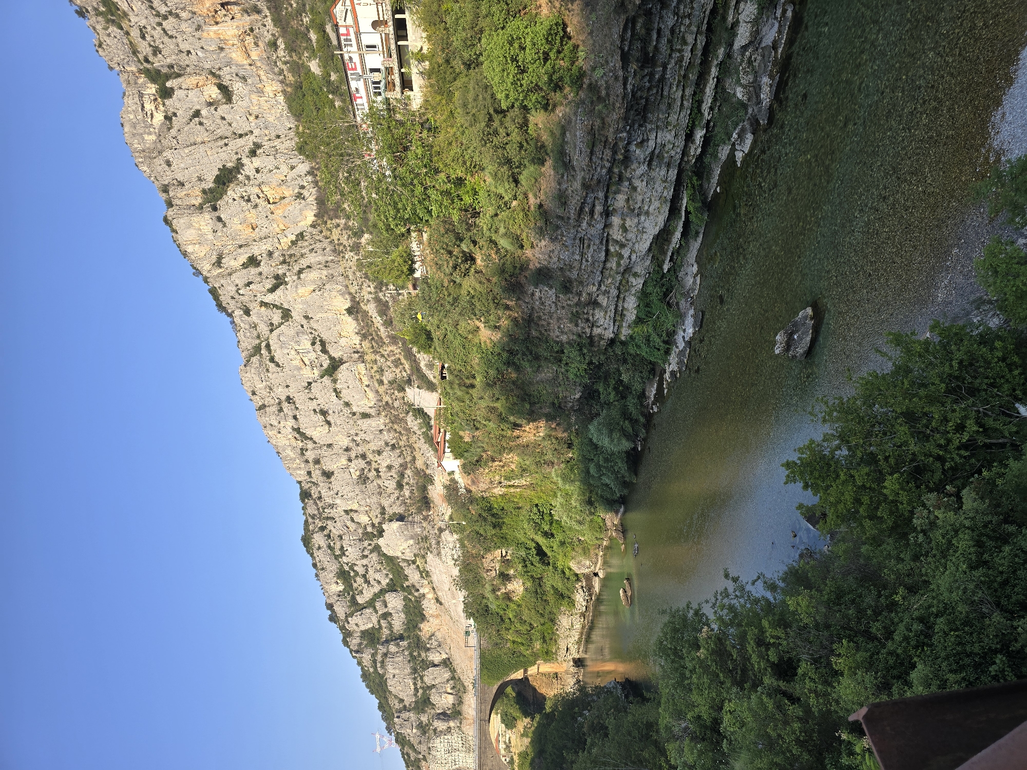 The image shows a wide, clear river flowing through a canyon. The riverbanks are lined with lush green trees and steep, rocky cliffs. A stone bridge with an arch crosses the river in the middle ground. On the right bank, several buildings are visible, nestled amongst the trees on the cliff face. The sky above is a clear, bright blue. The overall scene is serene and picturesque, showcasing a natural landscape with some human infrastructure.