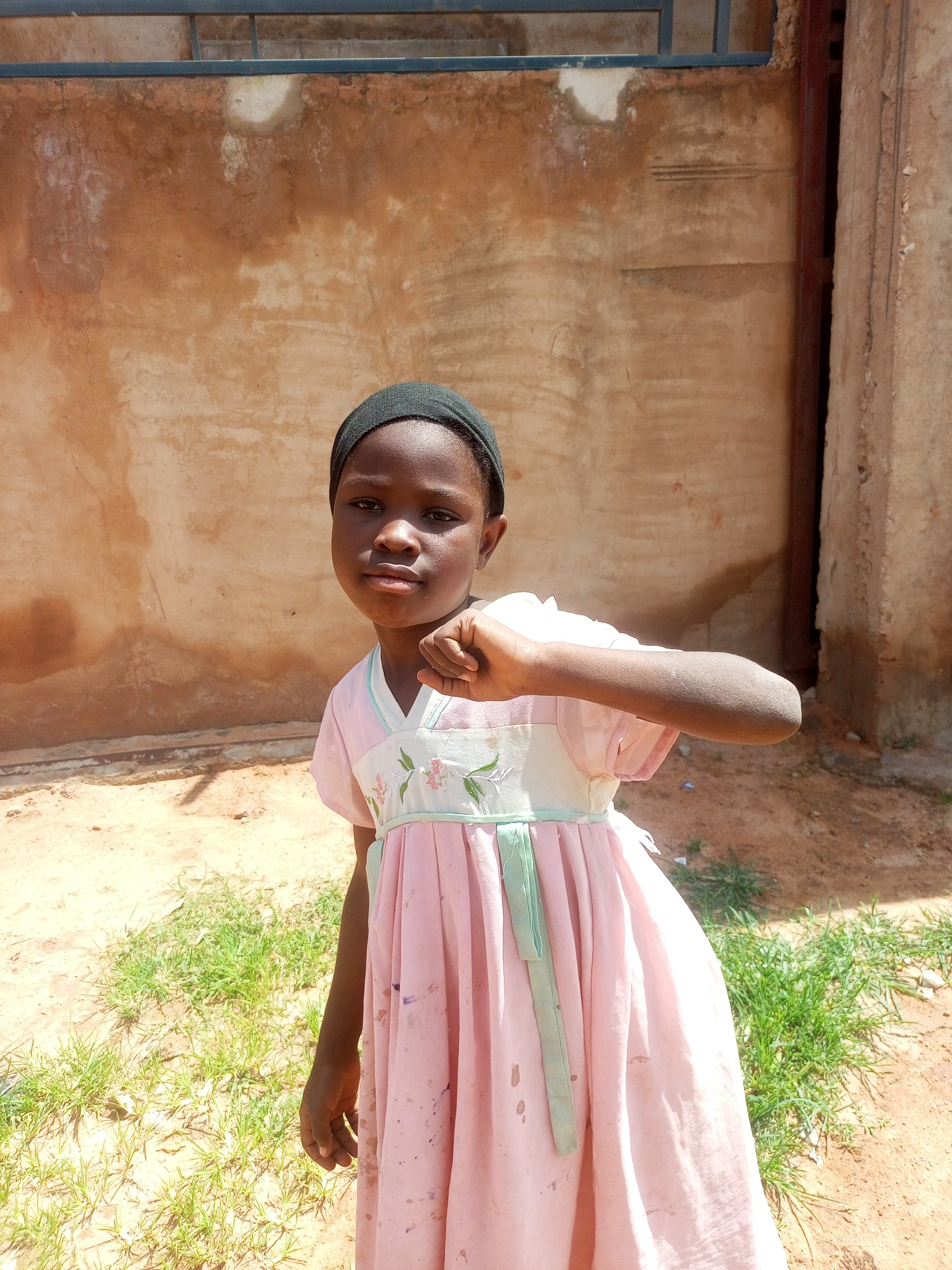 A young girl wearing a pink dress and a black head covering stands in front of a textured wall in Tempure, Nigeria. The wall, painted an earthy orange, shows signs of wear and has horizontal markings near the top. To the right, a portion of a rough, concrete pillar is visible. The ground in front of the wall is sandy and scattered with patches of green grass. The girl faces the camera, holding her right fist near her chest. Her dress features a light-colored bodice with embroidered floral patterns and a light green sash. The overall lighting suggests it is daytime.