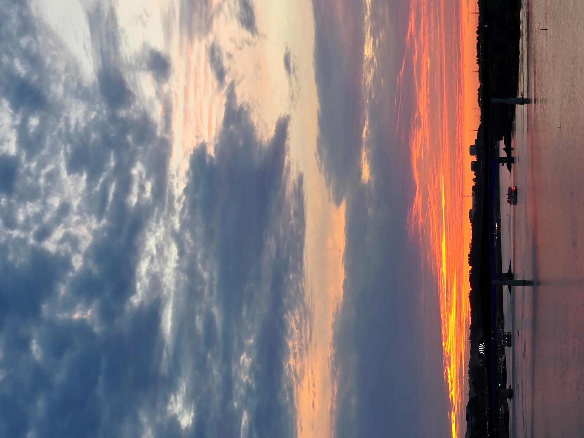 The image depicts a sunset over the Vistula River in Warsaw, Poland. A bridge, possibly the Siekierkowski Bridge, spans the river, with several boats visible on the water below. The sky is filled with dramatic clouds, transitioning from dark blues and grays at the top to vibrant oranges and reds near the horizon. Buildings and silhouetted trees line the riverbanks. The water reflects the colors of the sky, creating a shimmering surface.