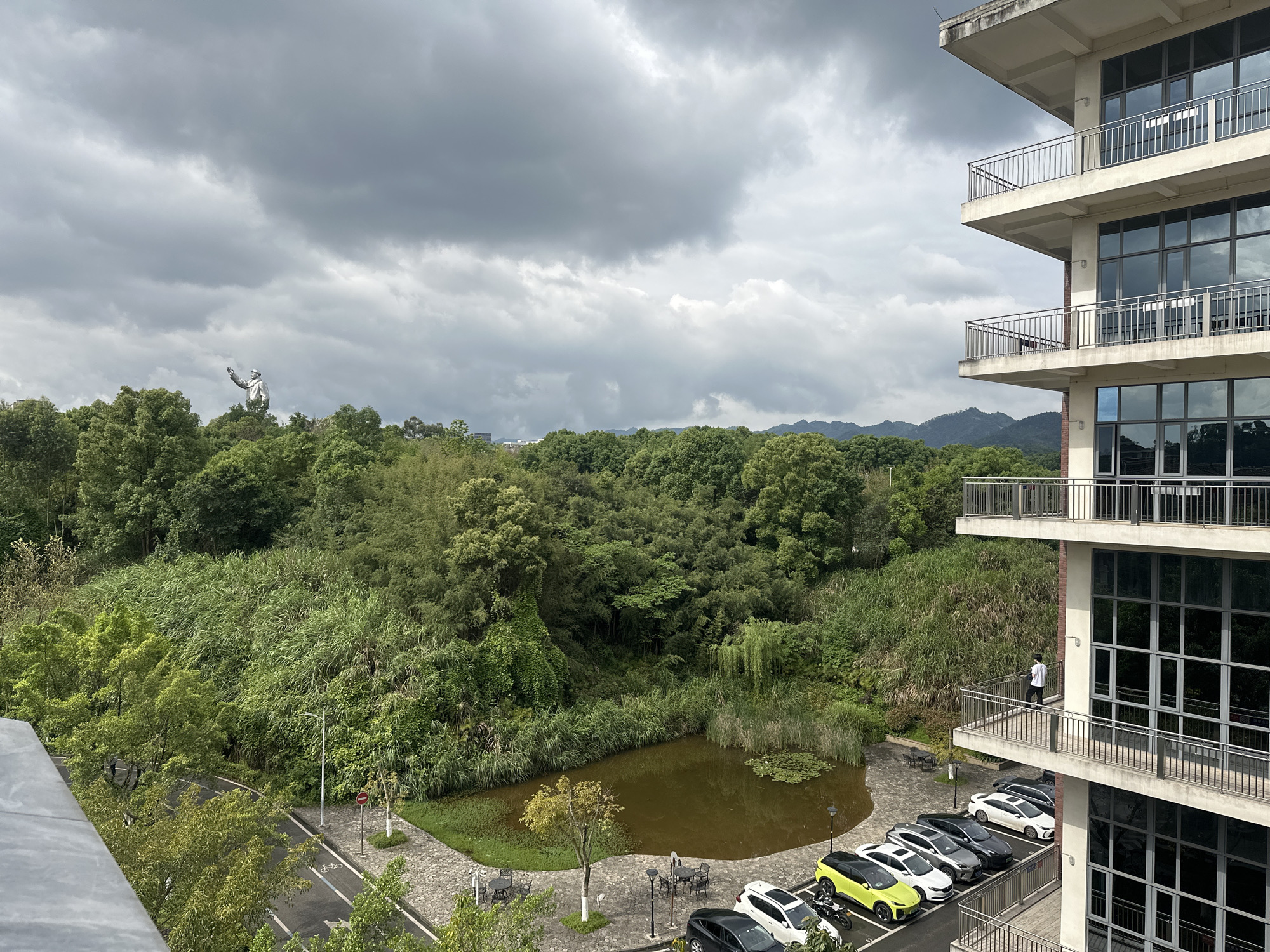 The image captures an elevated view of a mixed urban and natural landscape in what is identified as Chongqing High-tech Zone, China, under a heavily overcast sky.
The scene is dominated by dense, lush green vegetation covering hills and valleys in the midground and background, characteristic of a subtropical environment. A prominent, large, light-colored statue of a gesturing figure stands on a distant, tree-covered hill on the left side of the frame, partially obscured by the cloud-filled sky. In the center, nestled within the greenery, is an irregularly shaped pond with murky brown water and some water lilies, surrounded by tall reeds and bushes.
To the right, a multi-story, modern building with a light-colored exterior and brick accents extends vertically. It features multiple balconies with metal railings and large, reflective glass windows. A person in a white shirt and dark pants is standing on one of the lower balconies, looking outwards.
In the foreground and lower right, a paved area includes a parking lot with several parked cars of various models (SUVs and sedans, including a lime green SUV, white SUVs, and dark sedans) and a motorcycle. Adjacent to the parking lot is a small paved path with outdoor tables and chairs, and a paved road is visible further left. A white bicycle symbol is painted on the road, indicating a bicycle lane. A red octagonal stop sign with white text is visible near the road. Streetlights are also present. The distant background reveals faint outlines of mountains under the cloudy sky.
The weather is overcast, with thick grey clouds covering the sky, suggesting a cloudy or potentially rainy day, providing diffused lighting to the scene. The image captures a daytime setting.