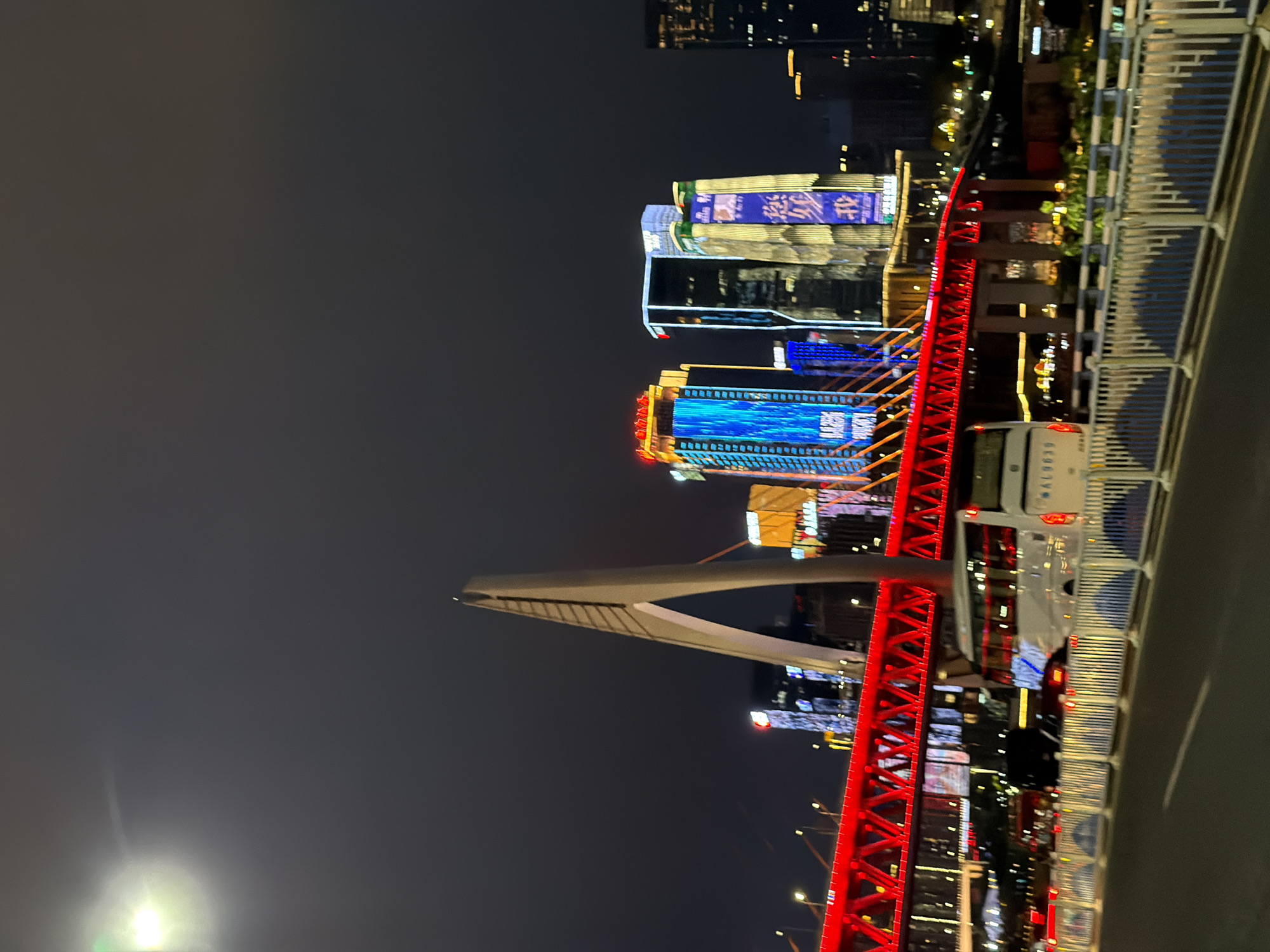 This is a nighttime street-level photograph of a modern cityscape with a prominent red illuminated bridge. The sky is dark and overcast. In the foreground, a multi-lane road is visible with a white and blue striped guardrail running along the side. A white bus with visible license plates "AL9839" is stopped or moving slowly on the road, positioned behind the guardrail. Beyond the road and guardrail, the red illuminated bridge spans across the scene, its intricate metal structure clearly visible and brightly lit. In the background, a collection of tall, modern buildings with illuminated facades rise against the dark sky. Some of these buildings display vibrant colored lights, primarily blue, yellow, and white, with some featuring large digital displays or signage. The overall impression is of a dynamic and well-lit urban environment at night. The image captures the Yuzhong District in Chongqing, China, known for its impressive architecture and illuminated bridges.