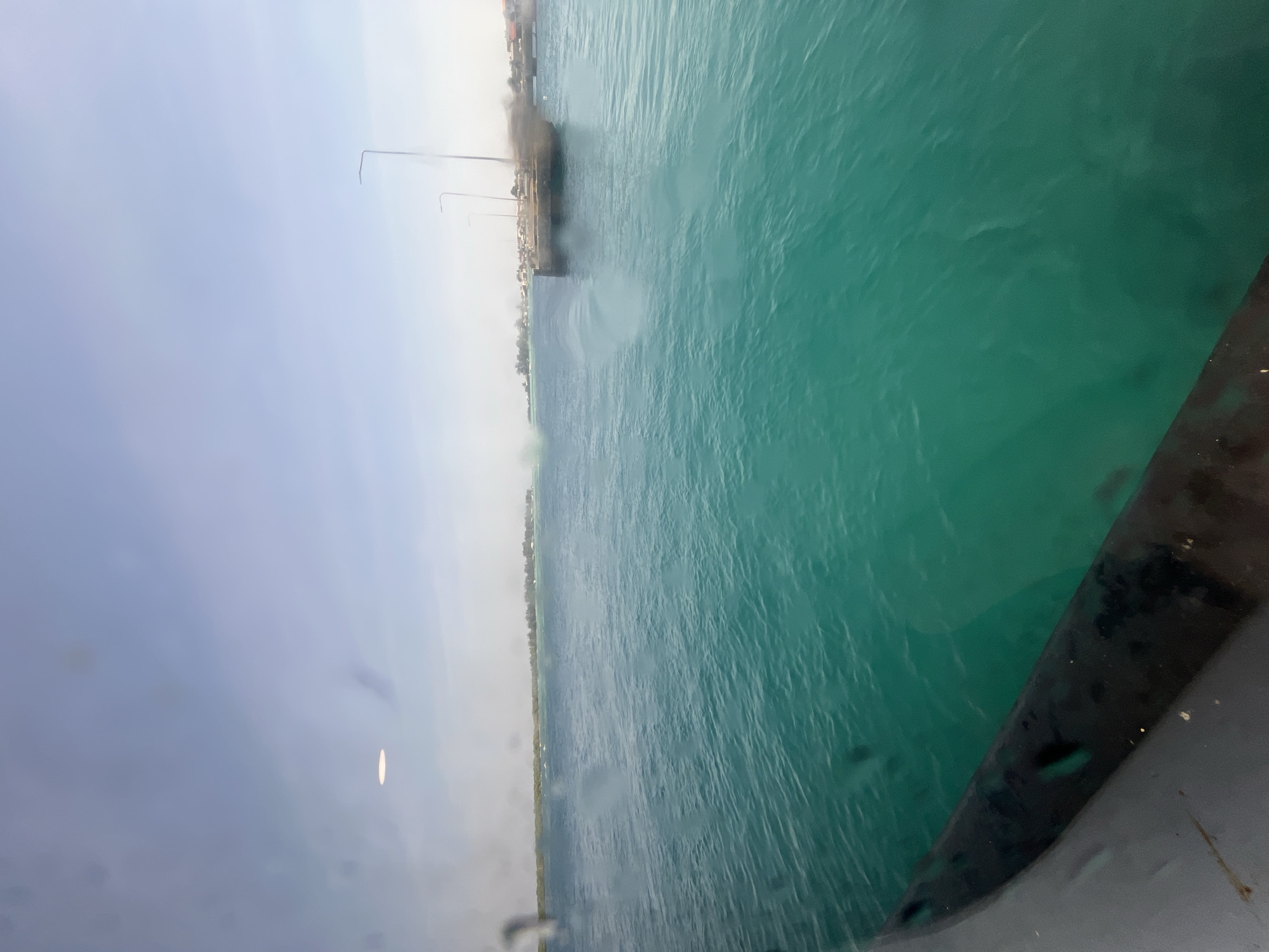 This image captures a view of the turquoise waters surrounding Karimunjawa, Indonesia. In the foreground, a dark, curved object, likely the railing of a boat, is visible. The water exhibits gentle ripples and appears clear, with the seabed possibly visible in some areas. In the distance, a narrow strip of land is lined with trees, suggesting the presence of islands or the coastline. A small harbor or dock area with buildings and streetlights can be seen on the right side of the image. The sky is a pale blue with some haze, indicating it might be daytime. The overall scene suggests a calm, tropical environment, characteristic of the Karimunjawa archipelago.