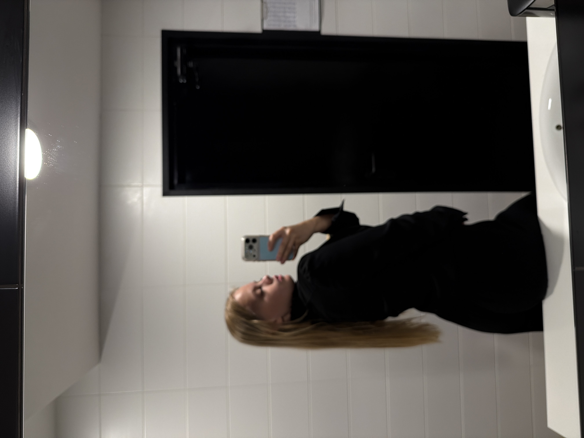 A young woman with long blonde hair takes a mirror selfie in what appears to be a public restroom. She is wearing a black blazer and is holding a light blue phone with a case. The background consists of white tiled walls and a black door. There is a white countertop in the foreground, and a sink is partially visible. The lighting suggests an indoor environment. No specific location in Izhevsk, Russia is identifiable from the image.