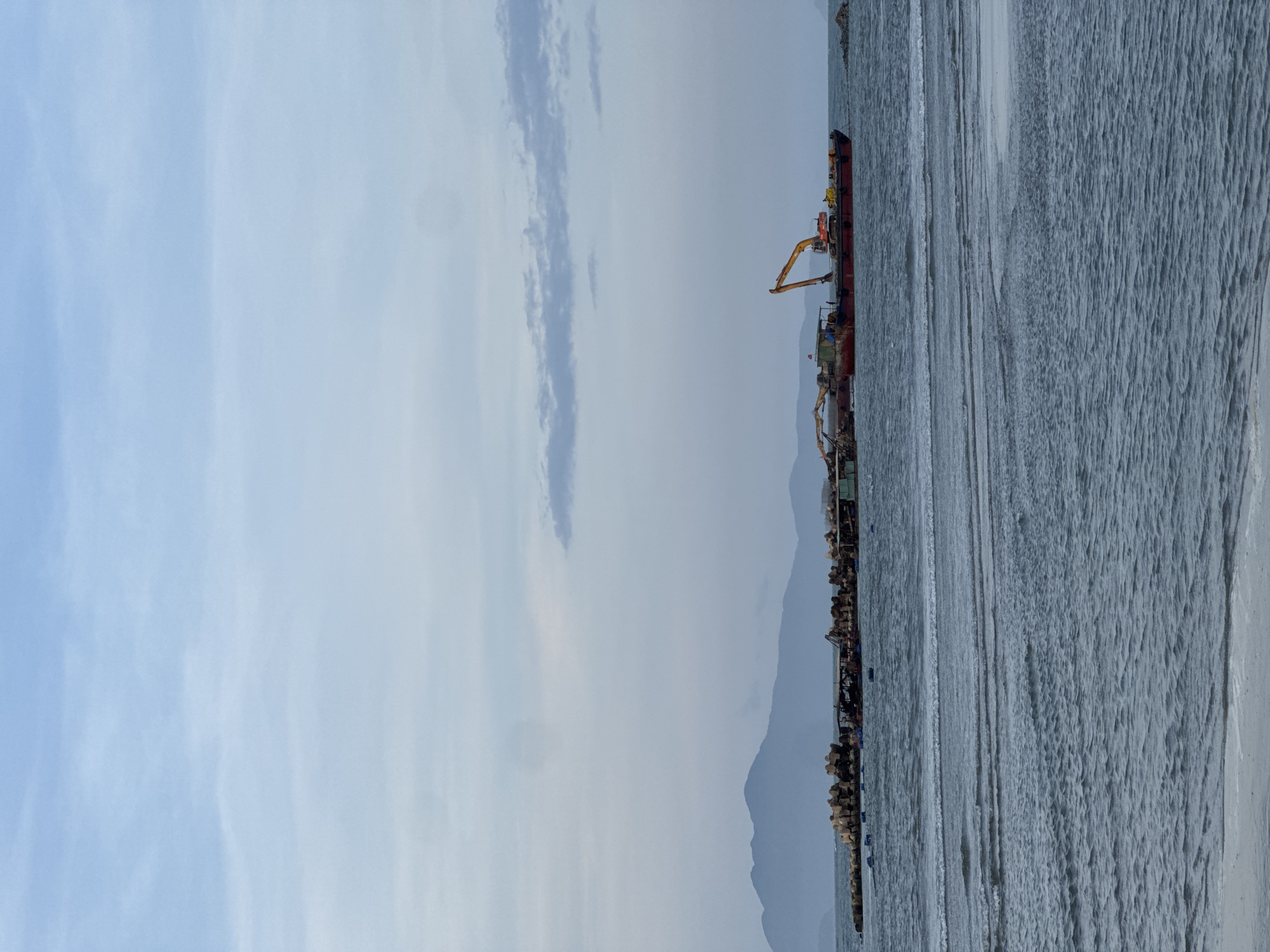 The image depicts a coastal scene in Đà Nẵng, Vietnam. A long pier or breakwater extends into the water, constructed from large, irregularly shaped rocks. An industrial barge with cranes and heavy equipment is docked along the structure. In the background, a mountain range is visible. The sky is overcast with scattered clouds. The water appears rough with visible waves.