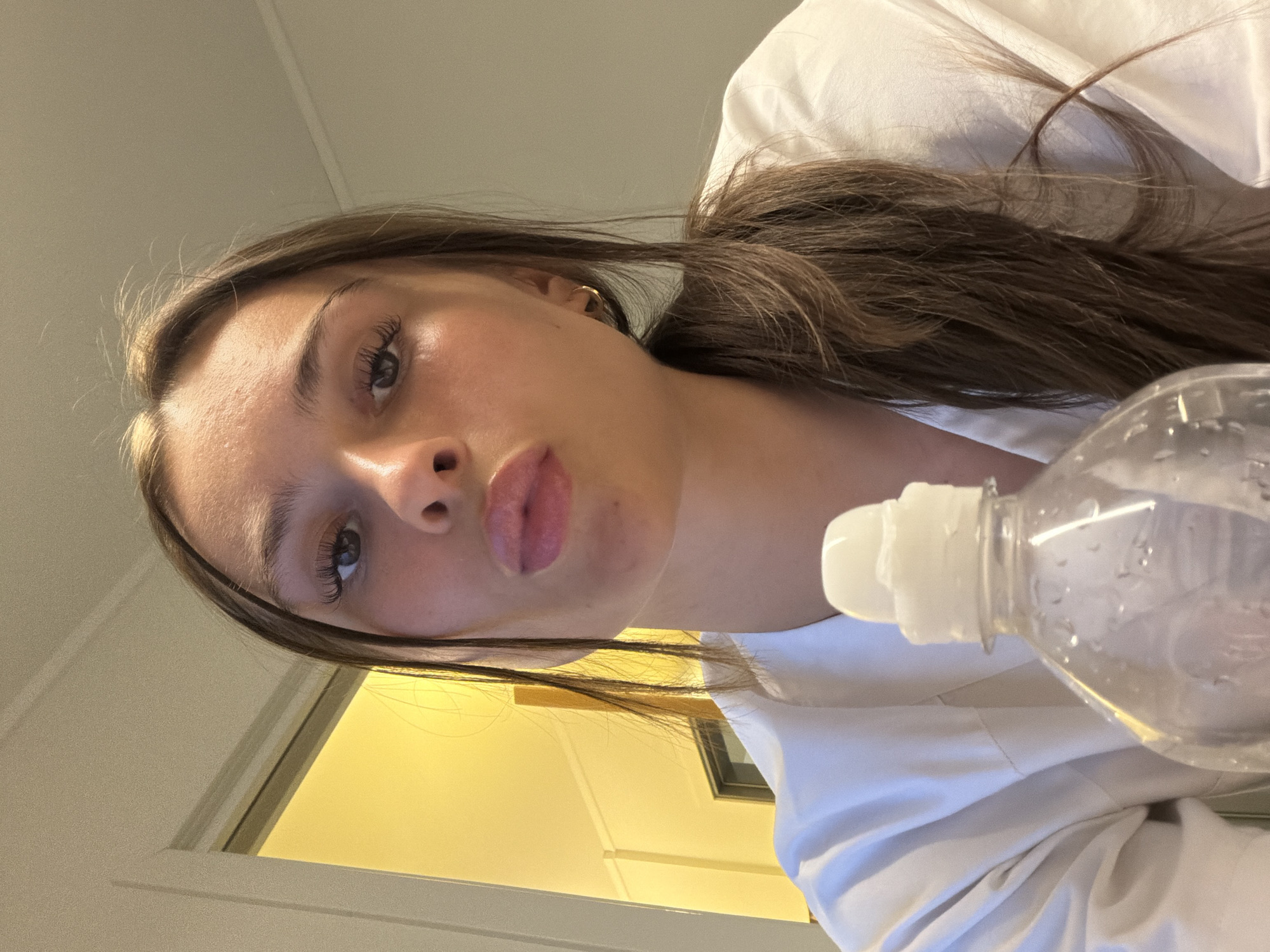A young woman with brown hair pulled back in a ponytail, wearing a white robe, poses for a selfie indoors. She has a water bottle in the foreground and is puckering her lips. The background shows a door frame with yellow light emitting from beyond it. There are no other people or animals visible in the image. The setting appears to be a residential interior, possibly a bathroom or bedroom, with neutral-colored walls and a door. The lighting is warm and artificial. There is no visible information to indicate the location as Harstad, Norway.