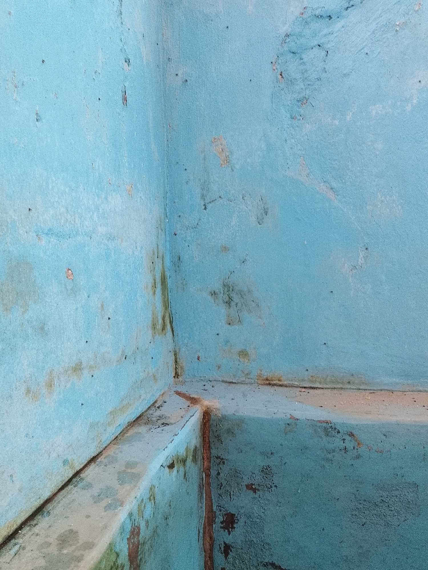 A close-up, eye-level photograph captures an interior corner of a blue-painted structure, likely a room or alcove, in Karangsari, Indonesia. The walls and a built-in concrete ledge are aged and show signs of wear. The blue paint is faded and peeling in places, revealing patches of lighter plaster or concrete underneath. Streaks of green and brown discoloration, possibly from water damage or mold, run down the walls, particularly along the corner and the edges of the ledge. Small, dark speckles are scattered across the surfaces, consistent with general wear and tear. The lighting appears to be ambient, casting no harsh shadows, suggesting an overcast day or an indoor setting with diffused light. There are no people or discernible objects other than the architectural elements themselves. The scene conveys a sense of disrepair and neglect.