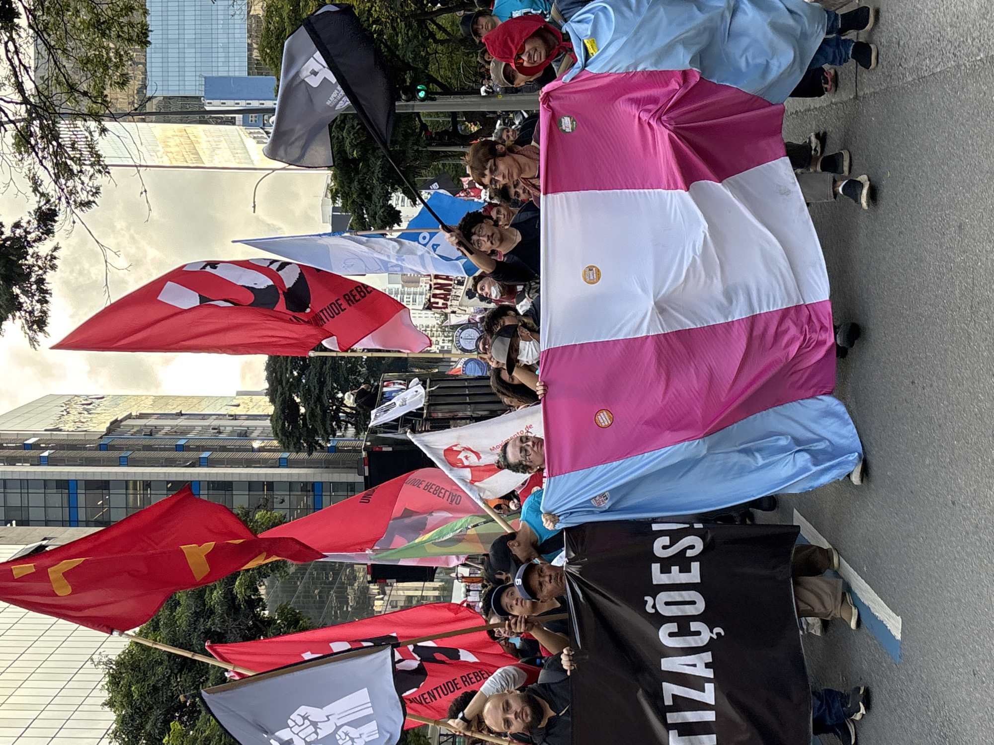 A crowd of people stand in the street, holding flags and banners, during what appears to be a protest or demonstration in the city of São Paulo, Brazil. Red flags are visible, and the black banner in the foreground bears the word "TIZAÇOES!" vertically. The backdrop features tall buildings, including modern glass structures, indicating an urban setting. The image appears to have been taken during daylight hours, as the sky is partly cloudy. The photo captures a sense of collective action and political expression.