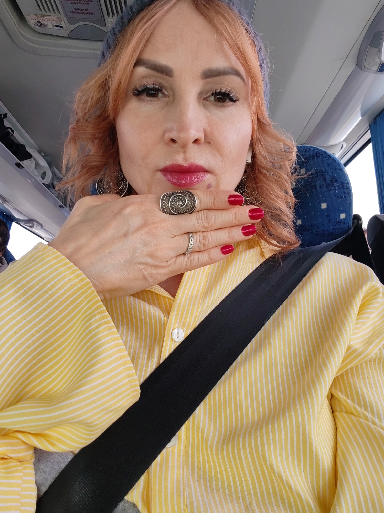 A person with reddish-orange curly hair and a blue beanie is seated on a bus with blue patterned seats and wearing a yellow and white striped shirt with a black seatbelt. They have red nail polish, are wearing two rings on their left hand, and have decorative earrings. The person is holding their hand to their chin. The inside of the bus is visible including overhead compartments and ceiling features. The location of Chaikovsky, Russia is not discernible from the image.