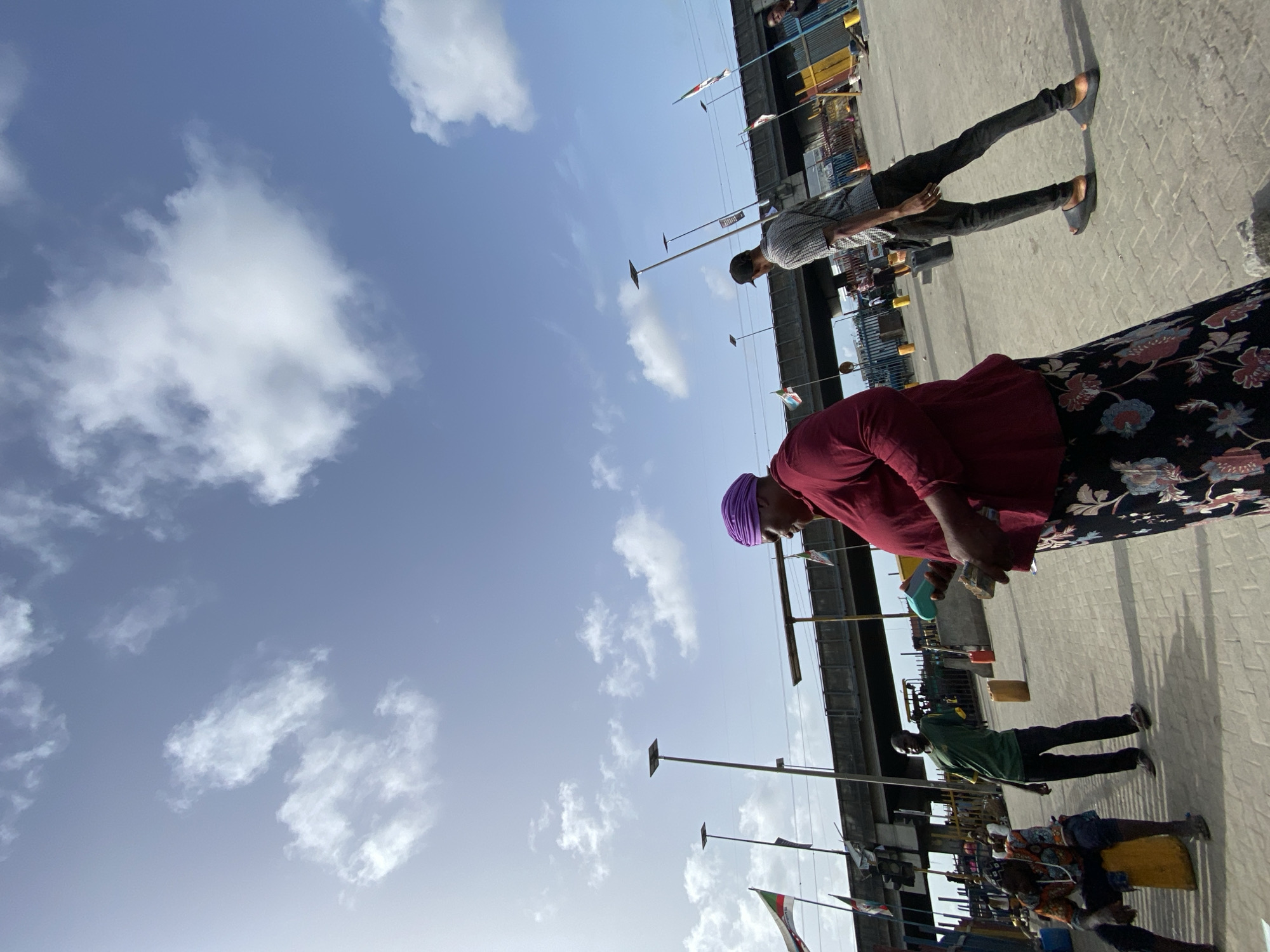 The image shows an outdoor scene in what appears to be a market area in Ado, Nigeria. The sky is light blue with scattered white clouds. A middle-aged woman wearing a purple headscarf, a red shirt, and a floral print skirt is the primary focus. She is holding objects in her hands, possibly money or goods. To her side, a man wearing a plaid shirt and dark pants is walking. In the background, there is an elevated structure, possibly a bridge or an overpass, decorated with numerous flags. Below the structure, other people are present, including one man wearing a green shirt and dark pants. The ground is paved with interlocking concrete pavers. Shadows indicate a strong light source, likely the sun. Overall, the image conveys the hustle and bustle of everyday life in a Nigerian market.
