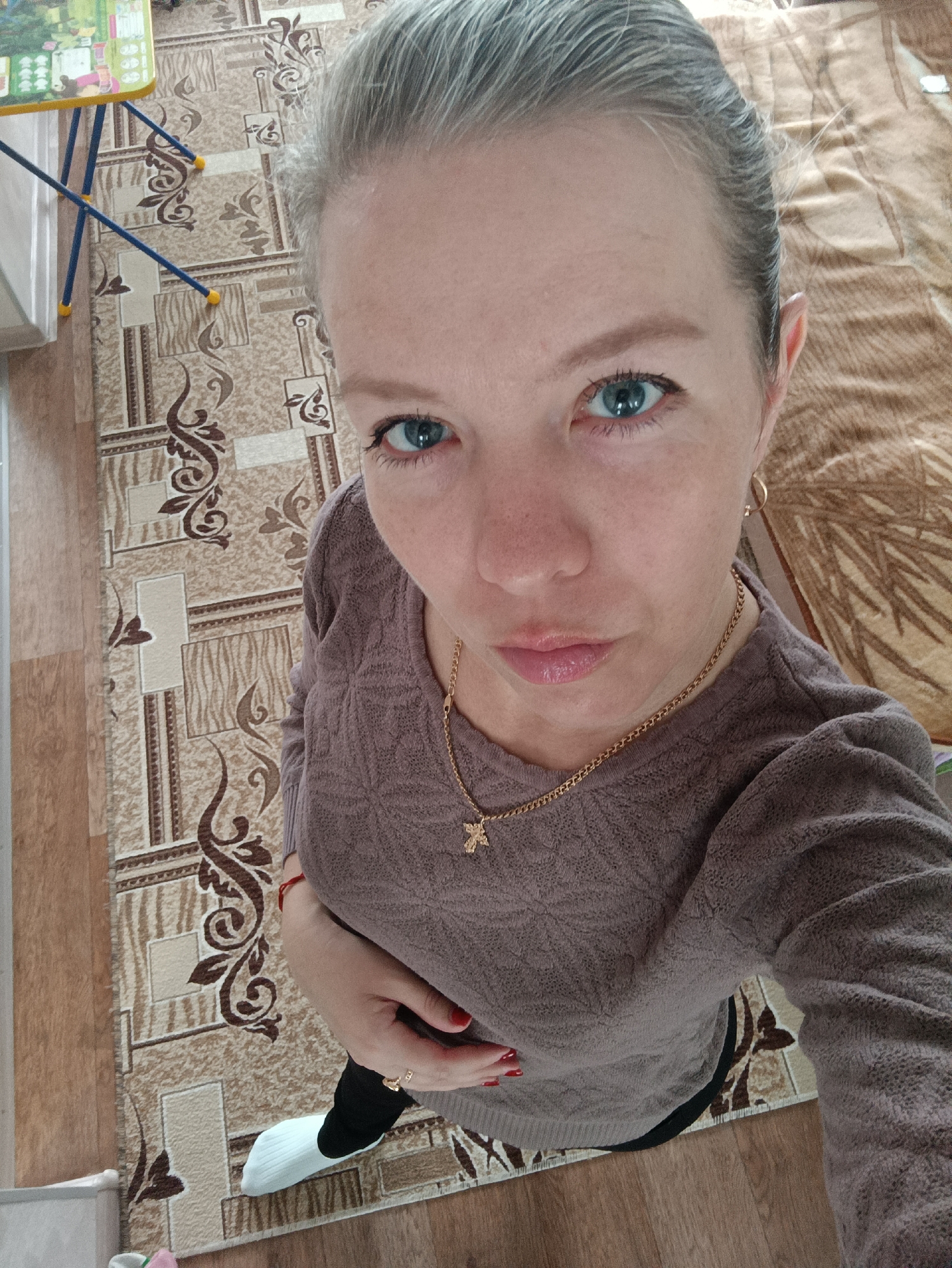 A fair-skinned woman with short, gray hair and blue eyes is looking directly at the camera in an indoor setting. She is wearing a brown long-sleeved sweater and a gold necklace with a cross pendant. Her fingernails are painted red. She is wearing black pants and a white sock. The background includes a patterned rug and a corner of what appears to be a child's table. The location is in Chaikovsky, Russia.