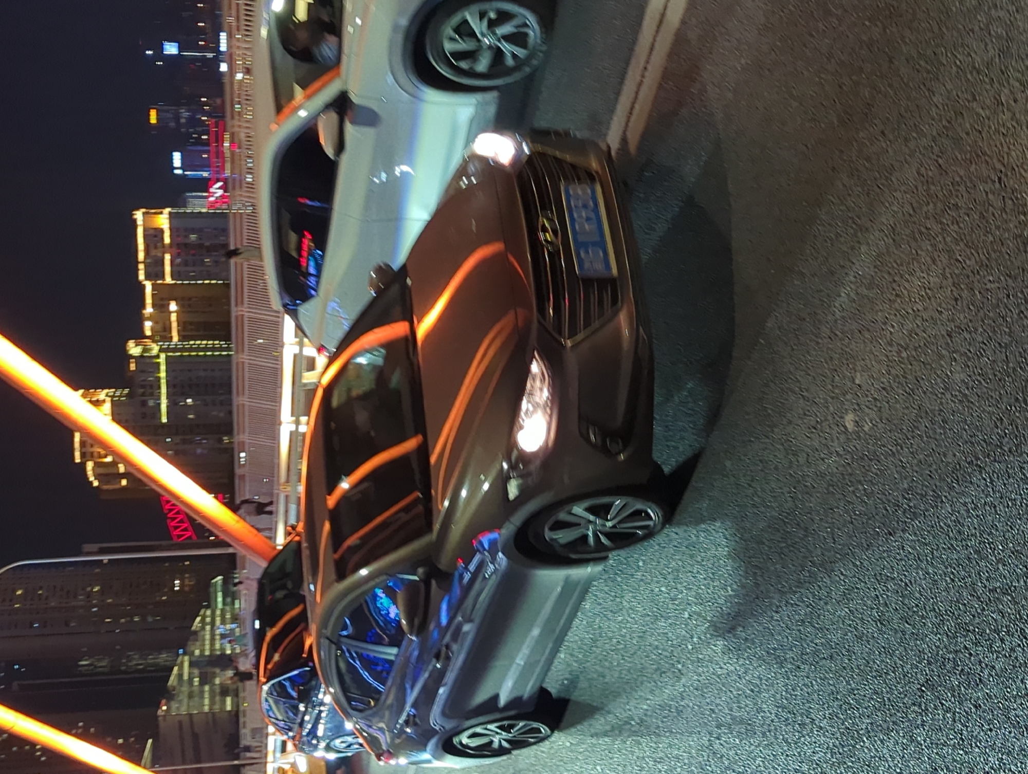 At night, on a paved road, a grey sedan is driving. The car is a Hyundai Elantra, and it has a license plate that reads "渝LR938". The car's headlights are on, and its wheels are spinning. In the background, there are tall buildings with lights on. There are also large, illuminated orange structures that appear to be part of a bridge or walkway. The overall scene suggests a busy urban environment at night.
