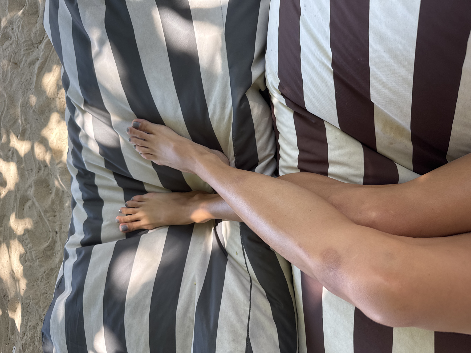 A pair of bare legs with grey nail polish on the toes rests on a striped cushion. The cushion has alternating stripes of dark grey and white. The legs are crossed at the knees. The background consists of sand with shadows from overhead foliage. The lighting suggests an outdoor setting, possibly a beach or a park, in Choeng Thale, Thailand.