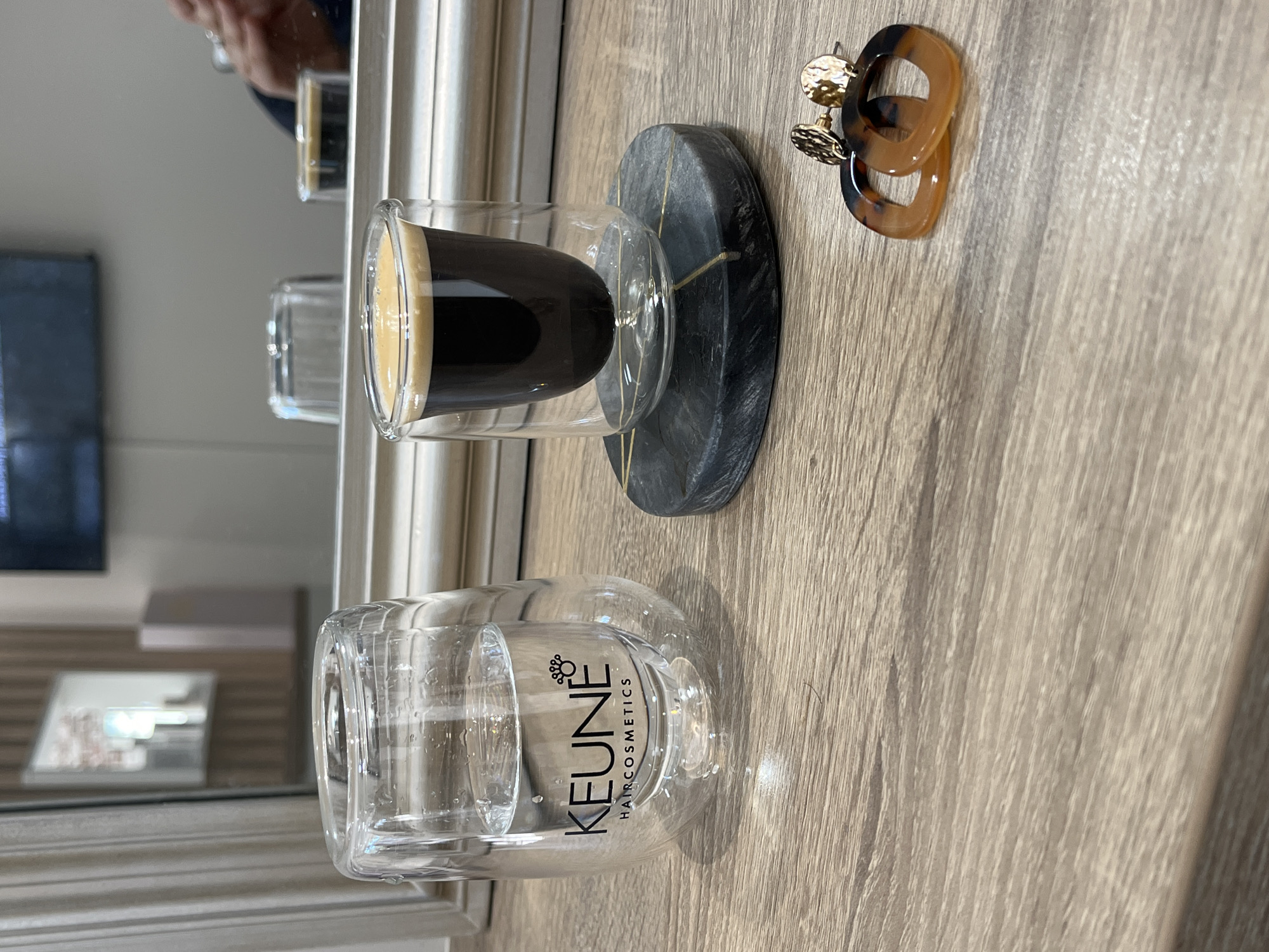 The image shows a close-up of a light-colored wooden surface. On the surface, there is a clear double-walled glass containing a dark liquid with a lighter-colored foam on top, possibly espresso. The glass is resting on a small, dark gray coaster with gold-colored veining. Next to the coaster are a pair of hoop earrings with a tortoise-shell pattern and gold-colored studs. In the background, there is a mirror reflecting the scene and revealing parts of a room with a TV screen on the wall and a framed picture. There is no specific information in the image to confirm the location in the city of Montrond-les-Bains, France.