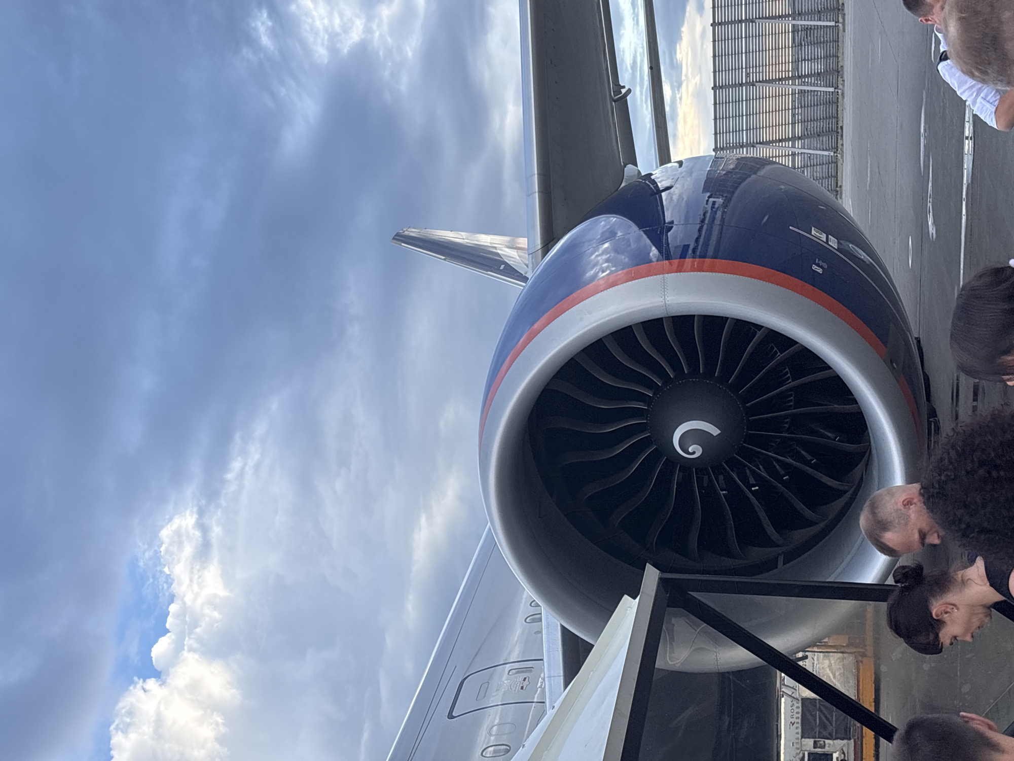 The image shows the engine of an airplane parked on an tarmac, likely at an airport. The sky is partly cloudy and the lighting suggests it is daytime, possibly late afternoon with the sun creating some warm hues in the distance. Several people are visible near the plane, some appearing to be passengers and others possibly airport personnel or guides. They are looking towards the aircraft, some with curiosity. The airplane's engine is large and prominent, with a dark turbine visible inside the intake. The fuselage is painted in dark blue with orange and white stripes. In the background, there is a metal structure, possibly part of the airport infrastructure or a hangar. One person in the foreground has curly black hair. Another person is wearing a light-colored shirt, possibly a uniform. The overall scene conveys a sense of arrival or departure at an airport. There are no clearly legible text elements in the image.