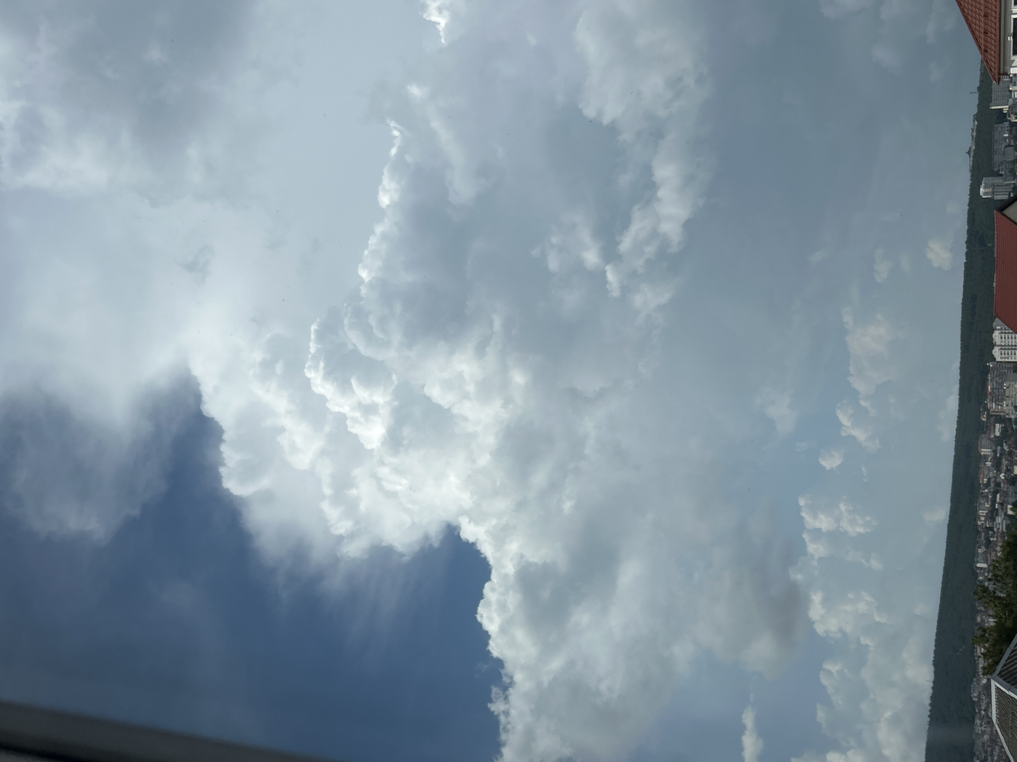 A view from a high vantage point looking out over the city of Gelendzhik, Russia, shows a vast sky filled with dramatic clouds. The clouds range from bright, fluffy white cumulus formations to darker, more ominous gray clouds. The lower portion of the image reveals the cityscape, with buildings and rooftops spread out below a dense line of green trees. The horizon is marked by a gentle slope covered in vegetation. The sky dominates the frame, suggesting a dynamic weather system.