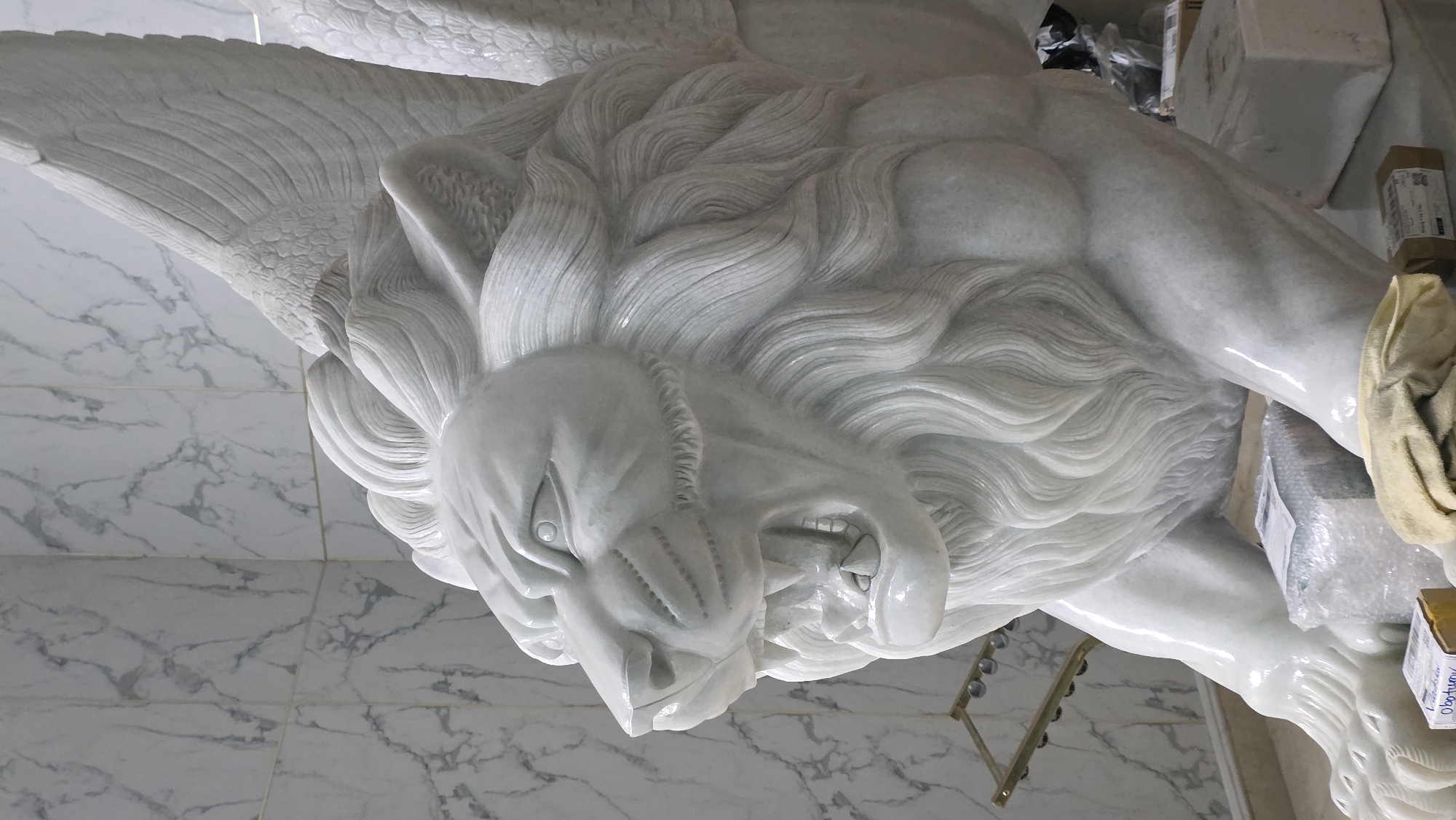 The image features a large, intricately carved white marble sculpture of a winged lion, positioned vertically, as if roaring. The lion's head, central to the view, displays a fierce expression with an open mouth, visible teeth, and an elaborate mane sculpted with flowing, wavy strands. Its eyes are deeply carved, enhancing its powerful gaze. Large, feathered wings extend from the lion's back, meticulously detailed with individual feathers. The entire sculpture is crafted from polished white marble, exhibiting a smooth, almost translucent finish with subtle grey veining.
The setting is an indoor space, likely a workshop, storage area, or showroom. The floor consists of large, light-colored marble tiles with prominent grey patterns. In the background, various objects are visible, including cardboard boxes, some wrapped in clear bubble wrap, and a yellow or tan cleaning cloth draped over an object to the right of the sculpture. A metallic structure with spherical components is partially seen on the floor. While no explicit activity is occurring, the presence of packaging and a cleaning cloth suggests the sculpture is either being prepared, unpacked, or maintained. Faint, partially obscured text, possibly "Obotunyo" or similar, is visible on a label on one of the boxes in the background. The overall impression is one of artistic craftsmanship and majestic power. The location context is Nha Trang Ward, Vietnam.