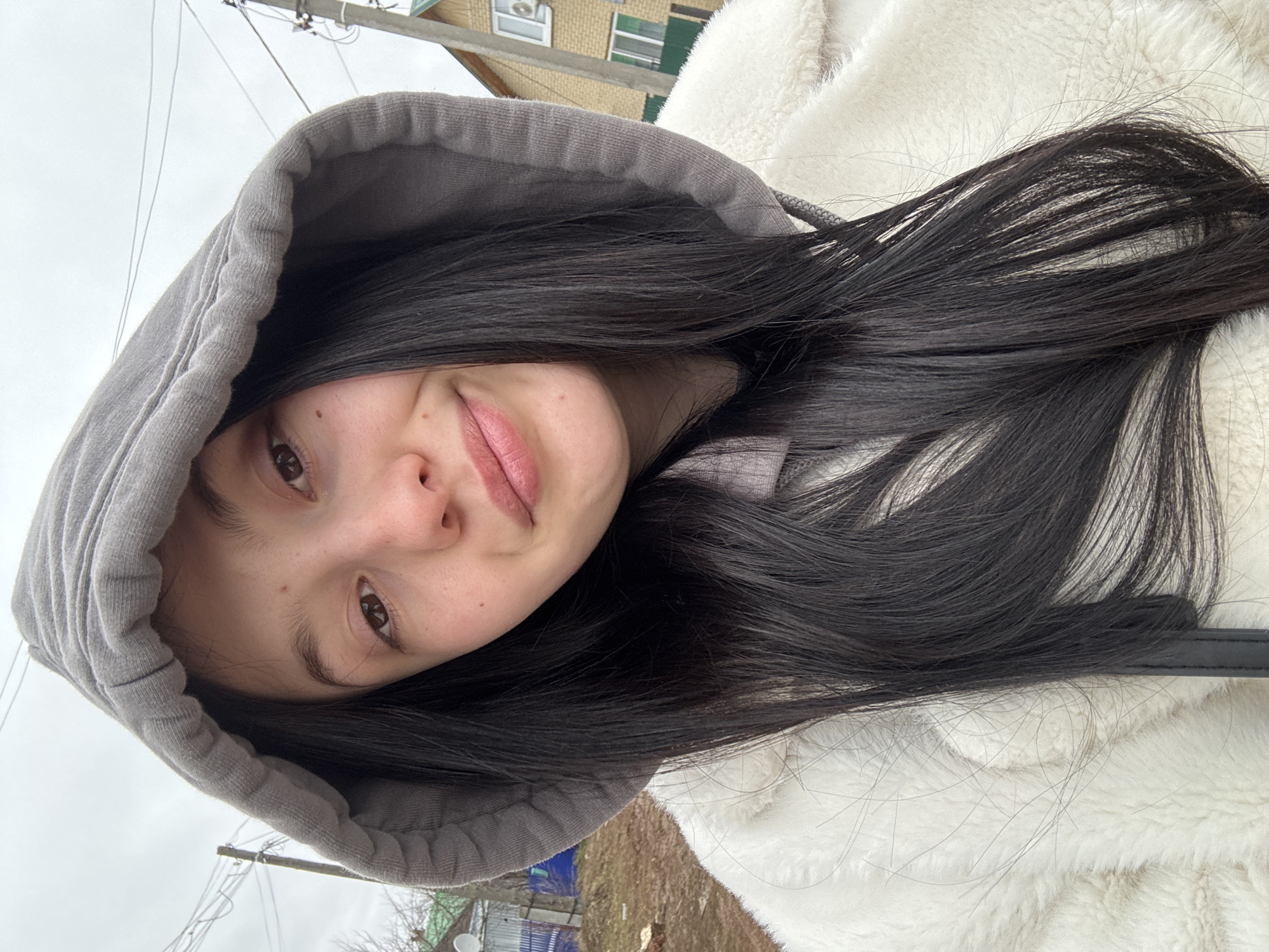 A young woman with long, dark hair is shown in a close-up, slightly rotated shot. She is wearing a gray hooded sweatshirt and a white, fluffy jacket. The location appears to be Фоки, Russia, evidenced by the background buildings and structures. The sky is overcast. There is a sense of being outdoors, likely in a residential area.
