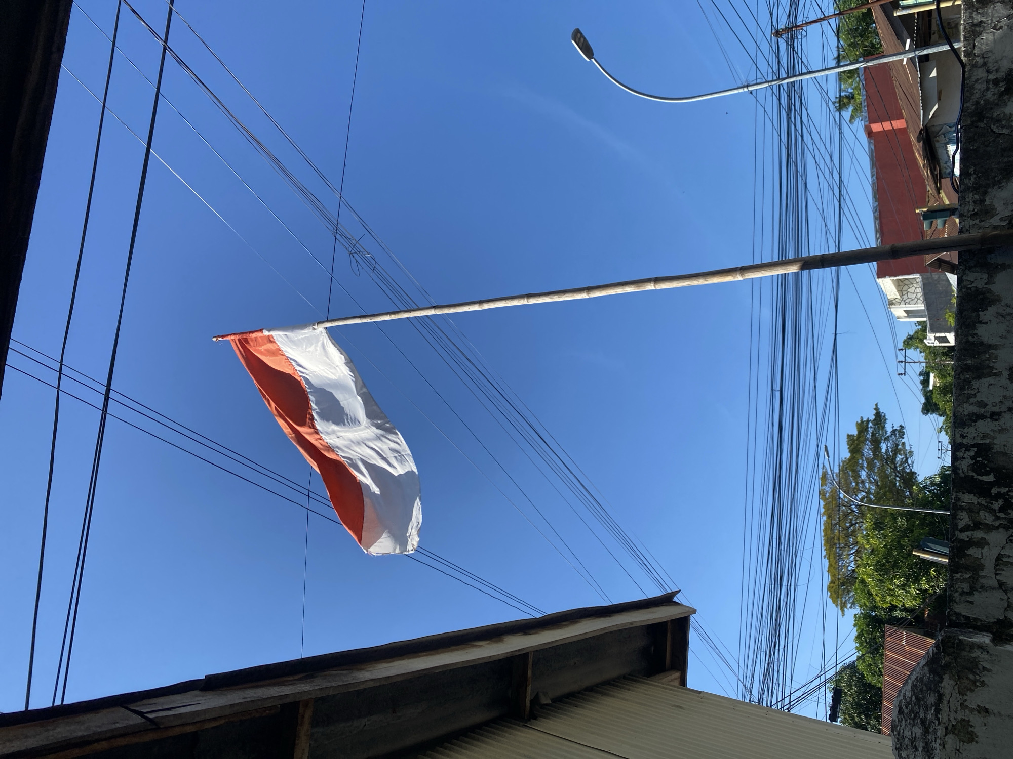 An Indonesian flag, with its red and white horizontal stripes, is raised on a bamboo flagpole against a clear blue sky. The flag appears to be waving in the wind. In the foreground, the edge of a building with a corrugated metal roof is visible on the left. A rough, concrete wall runs along the bottom of the image. Overhead, a dense network of electrical wires crisscrosses the sky. To the right, a modern street lamp with a curved pole stands against the blue backdrop. In the mid-ground, various buildings with tin roofs and some trees can be seen. The scene suggests an outdoor setting in a populated area, possibly a residential or commercial district in Bantaeng, Indonesia, given the presence of typical Indonesian architecture and infrastructure.