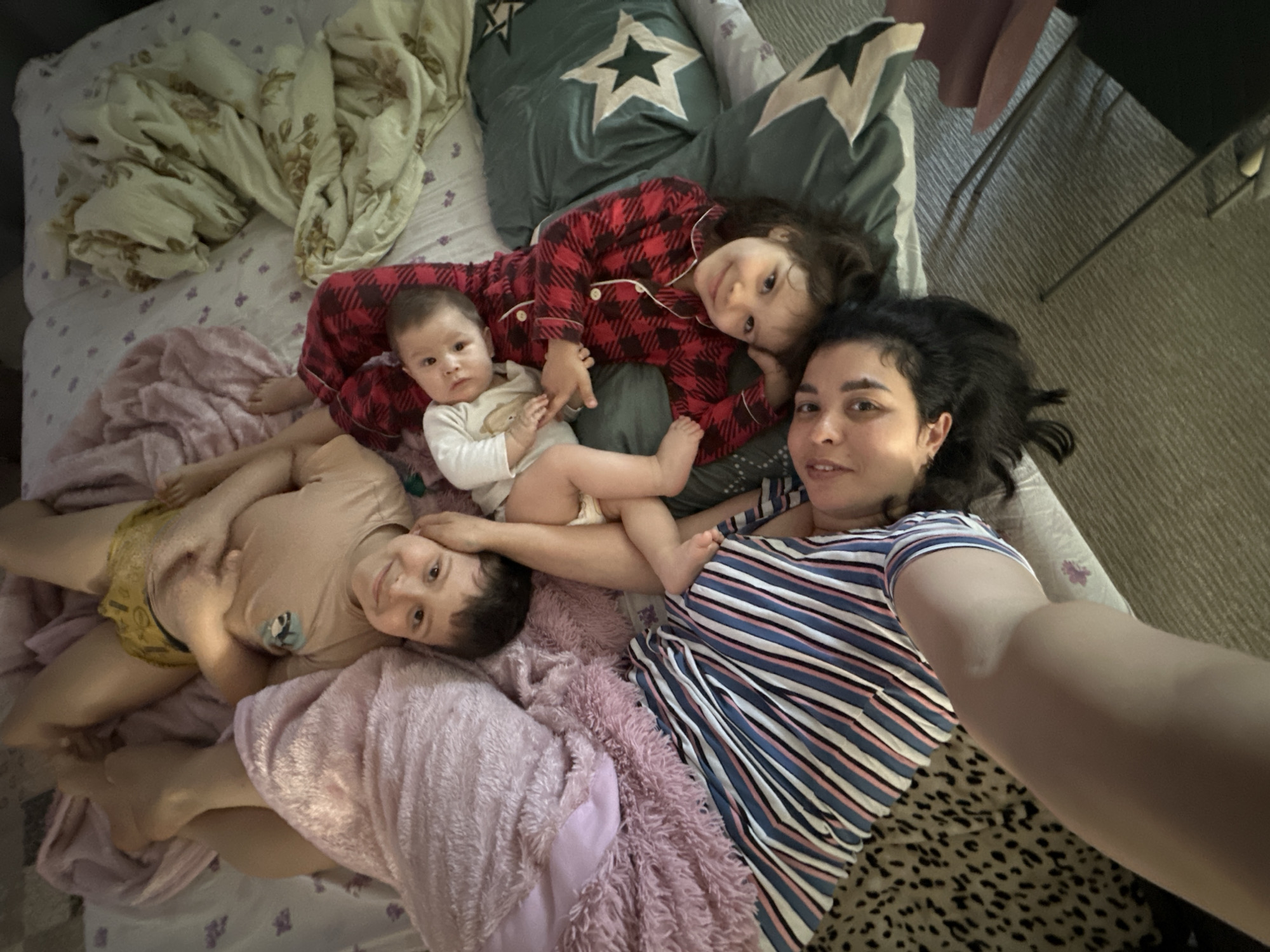 A selfie depicts a woman lying on a bed with three young children. The woman wears a blue and white striped shirt and has dark hair. One child, dressed in red and black plaid pajamas, is positioned near a pillow with a white star. Another child, wearing a white shirt and diaper, lies adjacent to the child in pajamas. A third child, clad in a beige shirt and yellow shorts, lies near a pink blanket. The bed is covered in patterned sheets and various blankets and pillows. A chair is visible in the background. The location is unknown.
