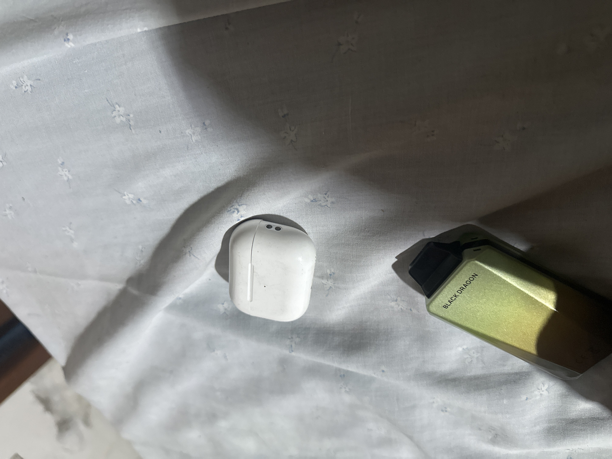The image displays two electronic devices situated on a white, textured fabric surface, with the scene located in the city of Juan Griego, Venezuela. In the upper middle of the frame, a white, plastic Apple AirPods Pro charging case is visible, exhibiting signs of wear and minor surface marks. Two small, dark, circular elements are discernible on its right side. Diagonally below the AirPods case, a metallic green and black vape device is present. The side of this device features the prominent text "BLACK DRAGON" in black capital letters. Both objects cast distinct, elongated shadows to their right and bottom, indicative of a single, strong light source originating from the upper left. The underlying fabric is a light-colored textile, possibly cotton, adorned with a repeating pattern of small, light blue floral or star-like motifs. A blurred, dark brown rectangular object is partially visible in the upper left background.