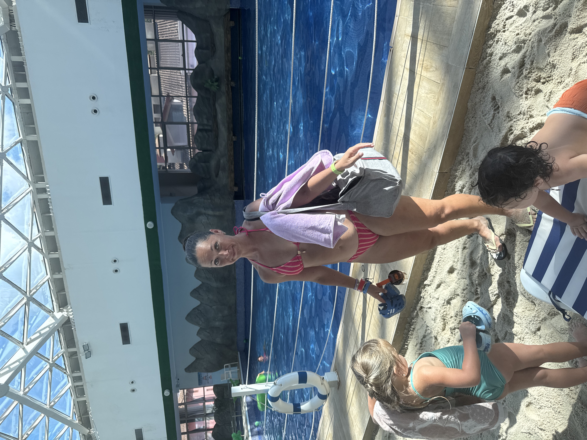 The media file depicts a group of people at an indoor swimming pool with a sandy area. A woman in a pink and white striped bikini is the central figure, carrying a light pink towel and a grey bag over her shoulder. She is standing on the sand next to the pool. In the foreground, a young girl in a teal swimsuit is sitting on the sand, holding a pair of blue sandals. Another child, partially visible, is also in the sandy area, wearing orange shorts and a blue and white striped garment.
The pool is a prominent feature, with clear blue water and visible lane markings. The background shows a rocky, artificial landscape, suggesting an indoor water park or themed resort. Large glass windows and a white ceiling with a geometric framework are visible, indicating an enclosed space. The lighting suggests it is daytime, with sunlight filtering through the windows. The sand area appears to be a designated relaxation zone. The overall mood seems relaxed and recreational. No visible text is present in the image. The location context is Esto-Sadok, Russia.