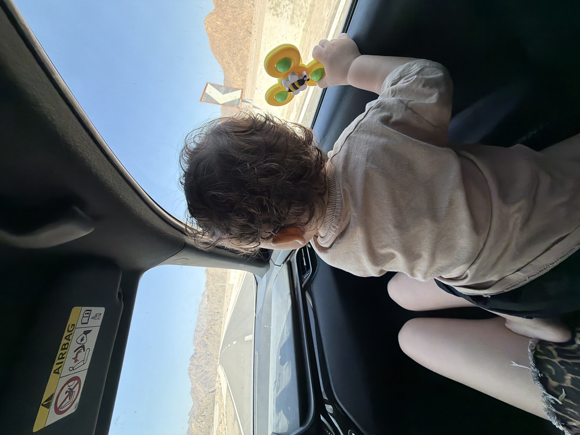 A toddler, seen from the rear, sits in the back seat of a car, looking out the window. The child has curly brown hair and wears a light-colored t-shirt and dark shorts. In their right hand, they hold a yellow and green toy bee. Beside the toddler, an adult's legs are visible, one bare and the other covered by a skirt with a leopard print pattern. The car's interior is dark, with the dashboard and a portion of the driver's side visible. The window reveals a desert landscape under a clear blue sky. A two-lane asphalt road stretches into the distance, flanked by rocky, arid hills. A road sign with a white chevron on an orange background is visible on the right side of the road. The overhead visor of the car displays an "AIRBAG" warning symbol and accompanying icons.